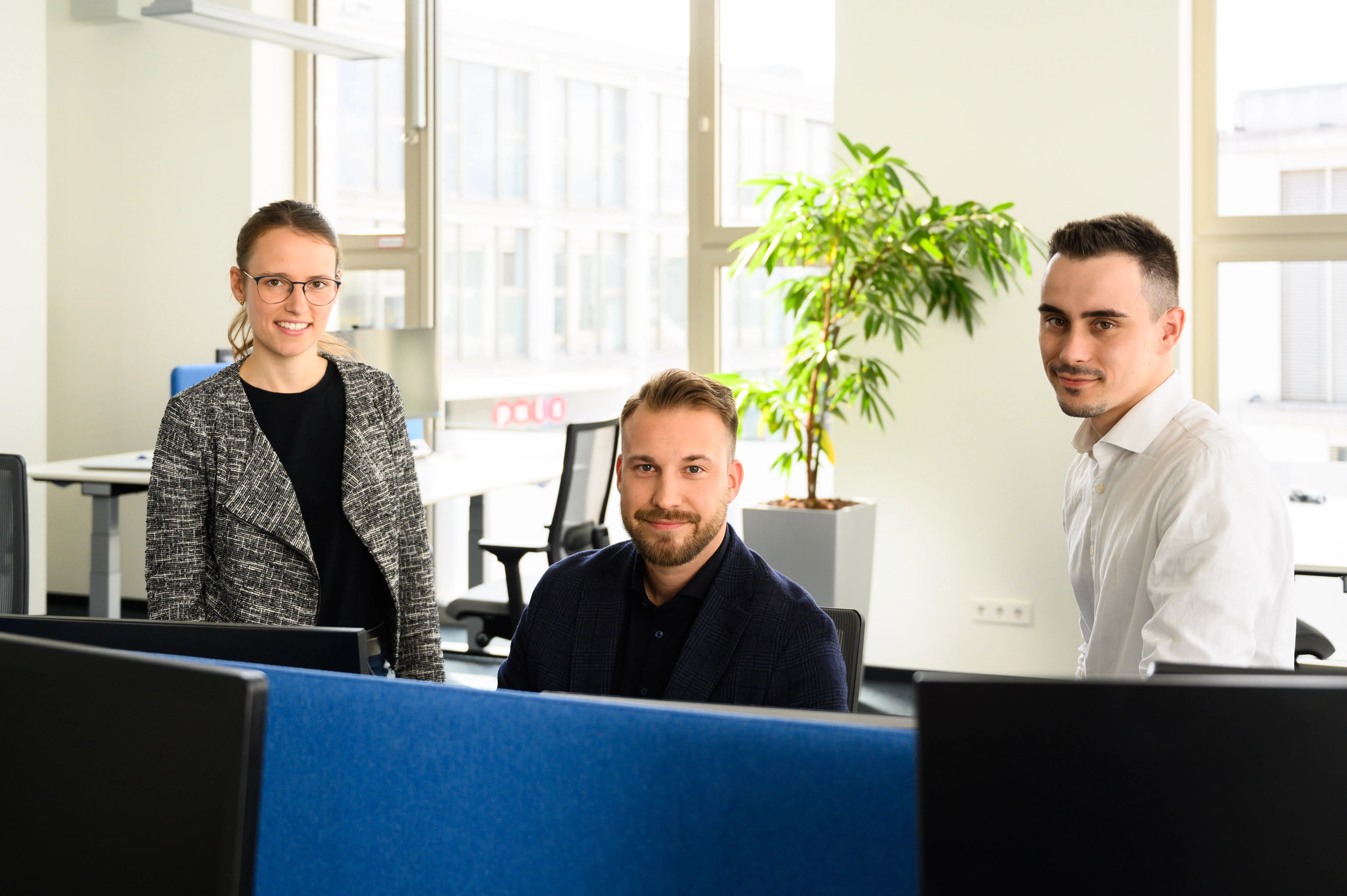 Drei IT-Fachleute in einem modernen Besprechungsraum mit Laptops, die in die Kamera lächeln.