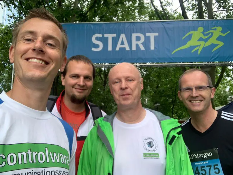 Vier Kollegen stehen lächelnd vor einem Startbanner bei einem Firmenlauf im Freien.
