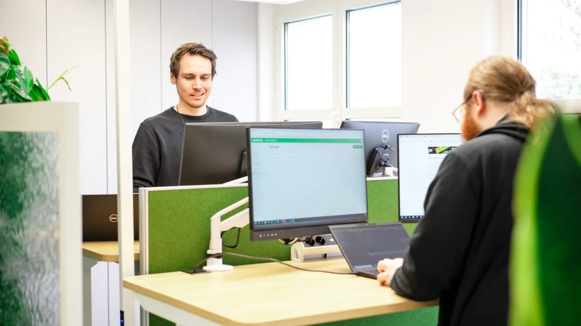 Zwei Kollegen arbeiten an höhenverstellbaren Schreibtischen im modernen, hellen Büro mit mehreren Monitoren.