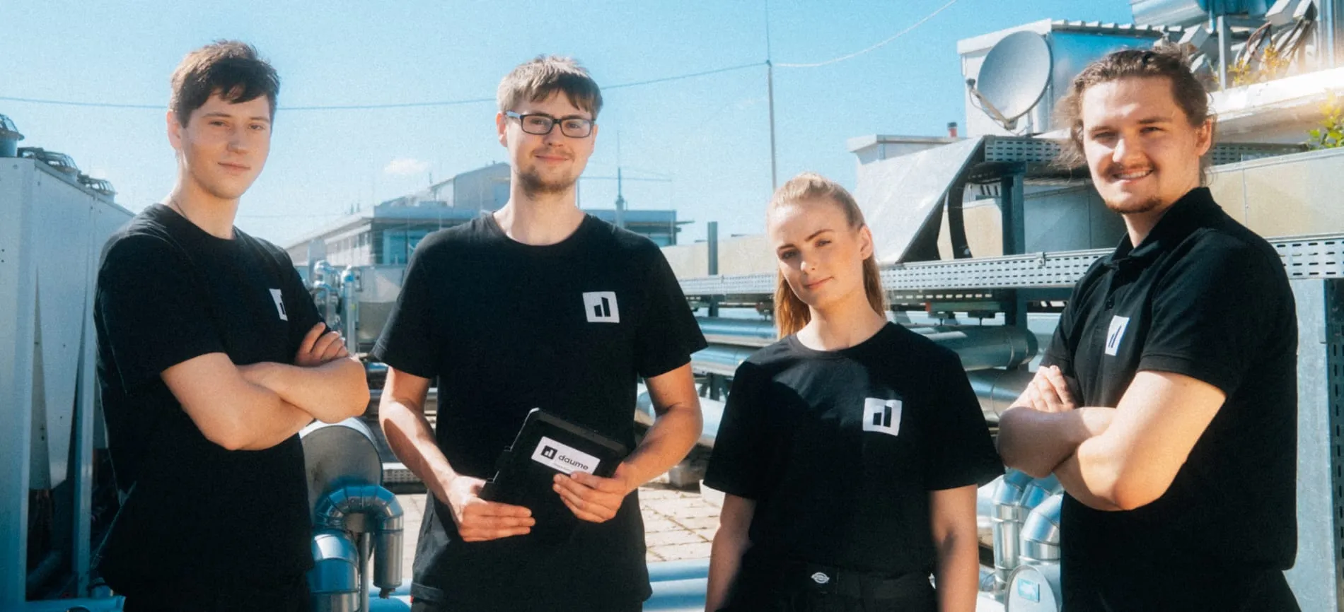 Vier junge Kollegen im Team-Outfit stehen gemeinsam auf einem Industriegelände und schauen freundlich in die Kamera.