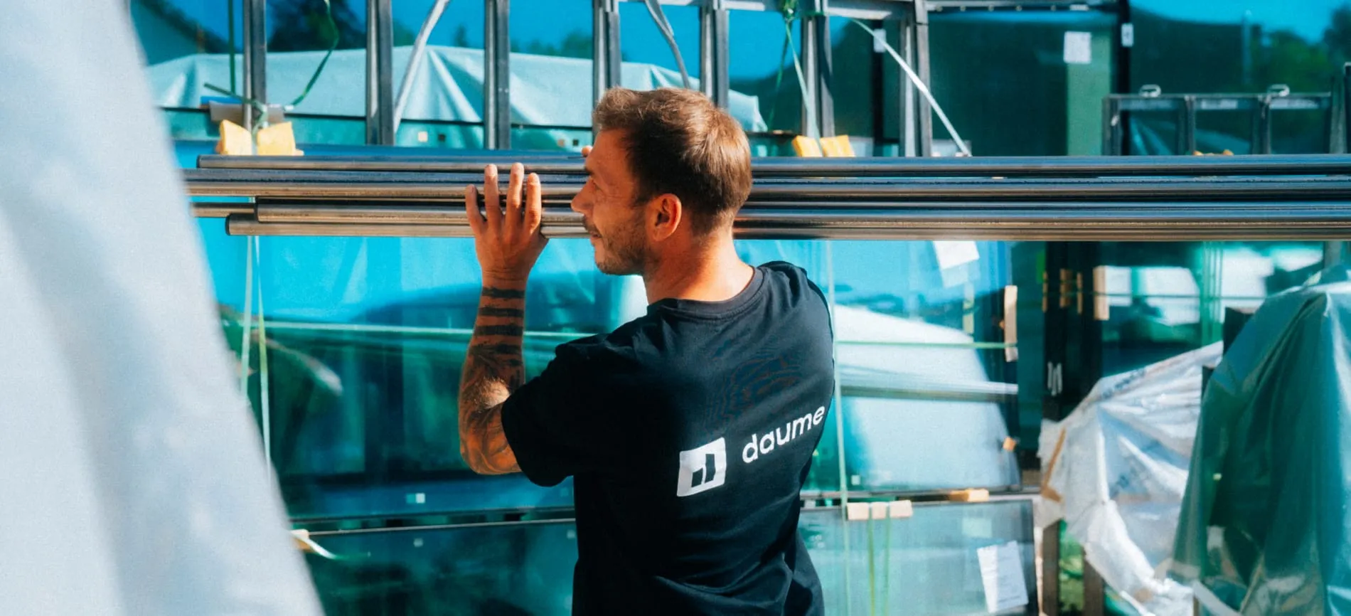 Du siehst einen Handwerker im Freien, der Metallrohre trägt und ein T-Shirt mit dem Logo "daume" trägt.