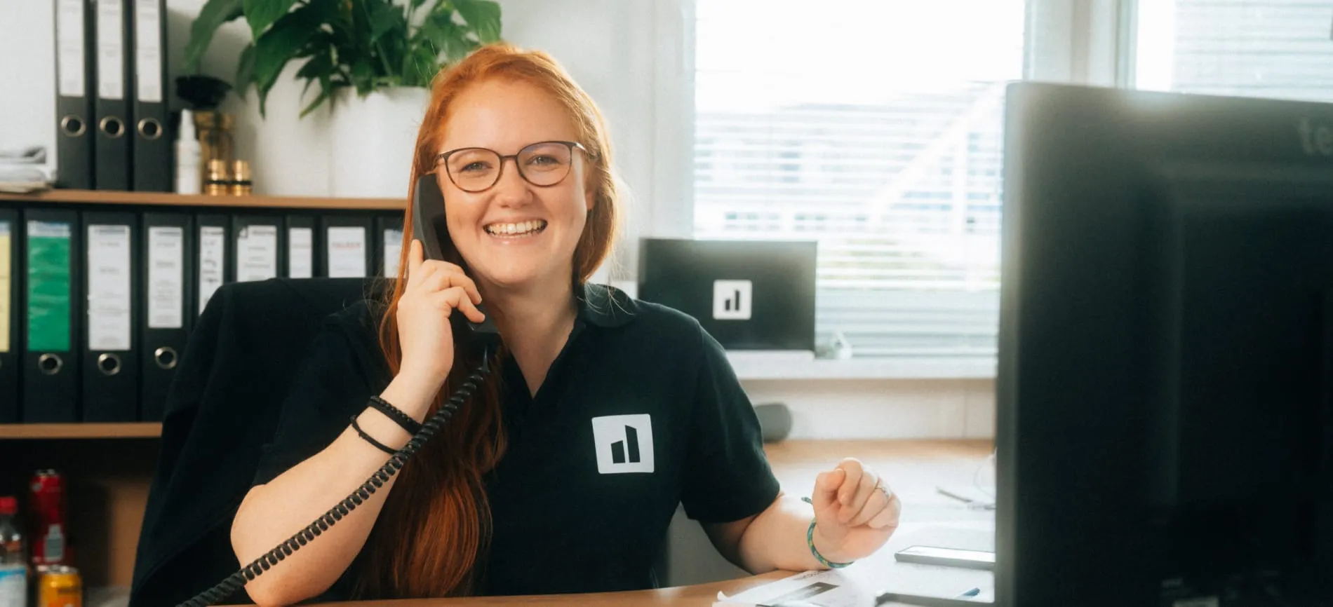 Du siehst eine lächelnde Mitarbeiterin im Büro, die am Schreibtisch telefoniert und dich freundlich begrüßt.