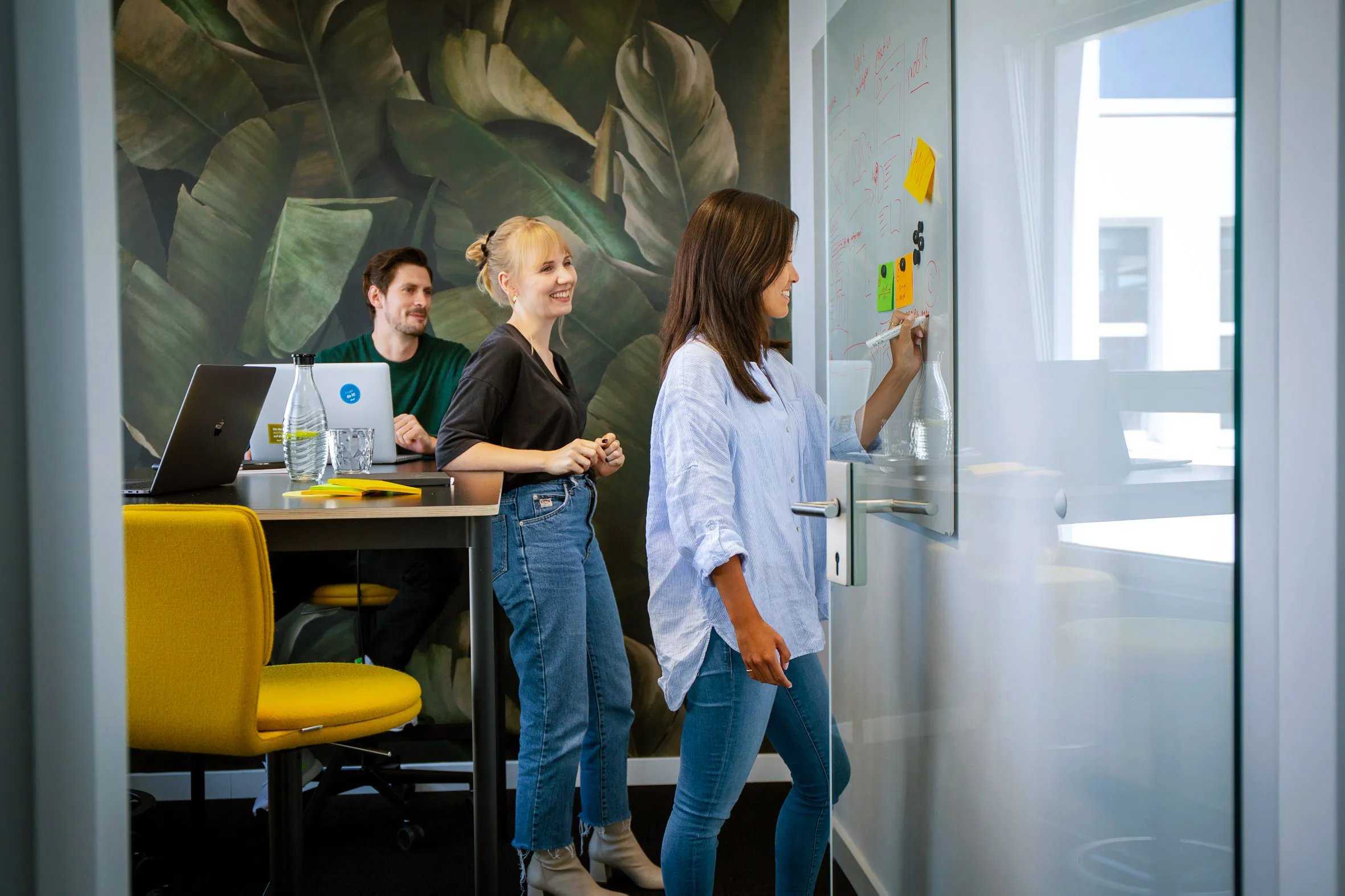 Drei Personen in einem modernen Meetingraum, diskutieren und schreiben auf ein Whiteboard.