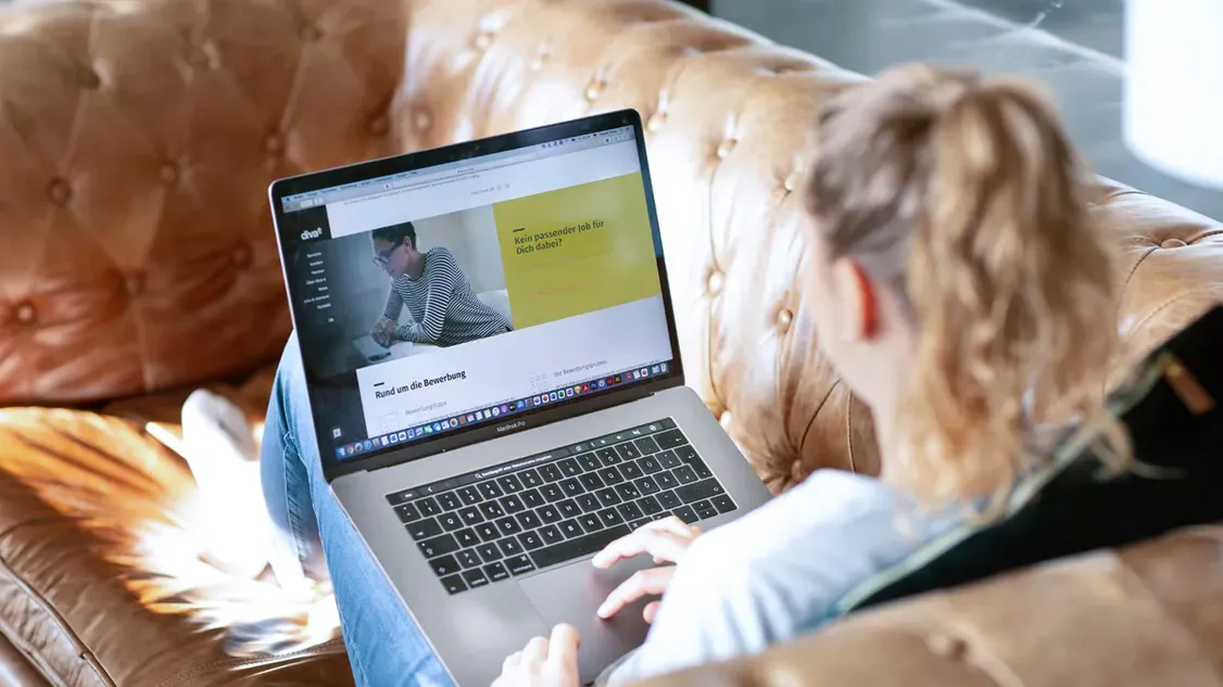 A woman is relaxing on a comfy couch, browsing the diva-e career site on a laptop.