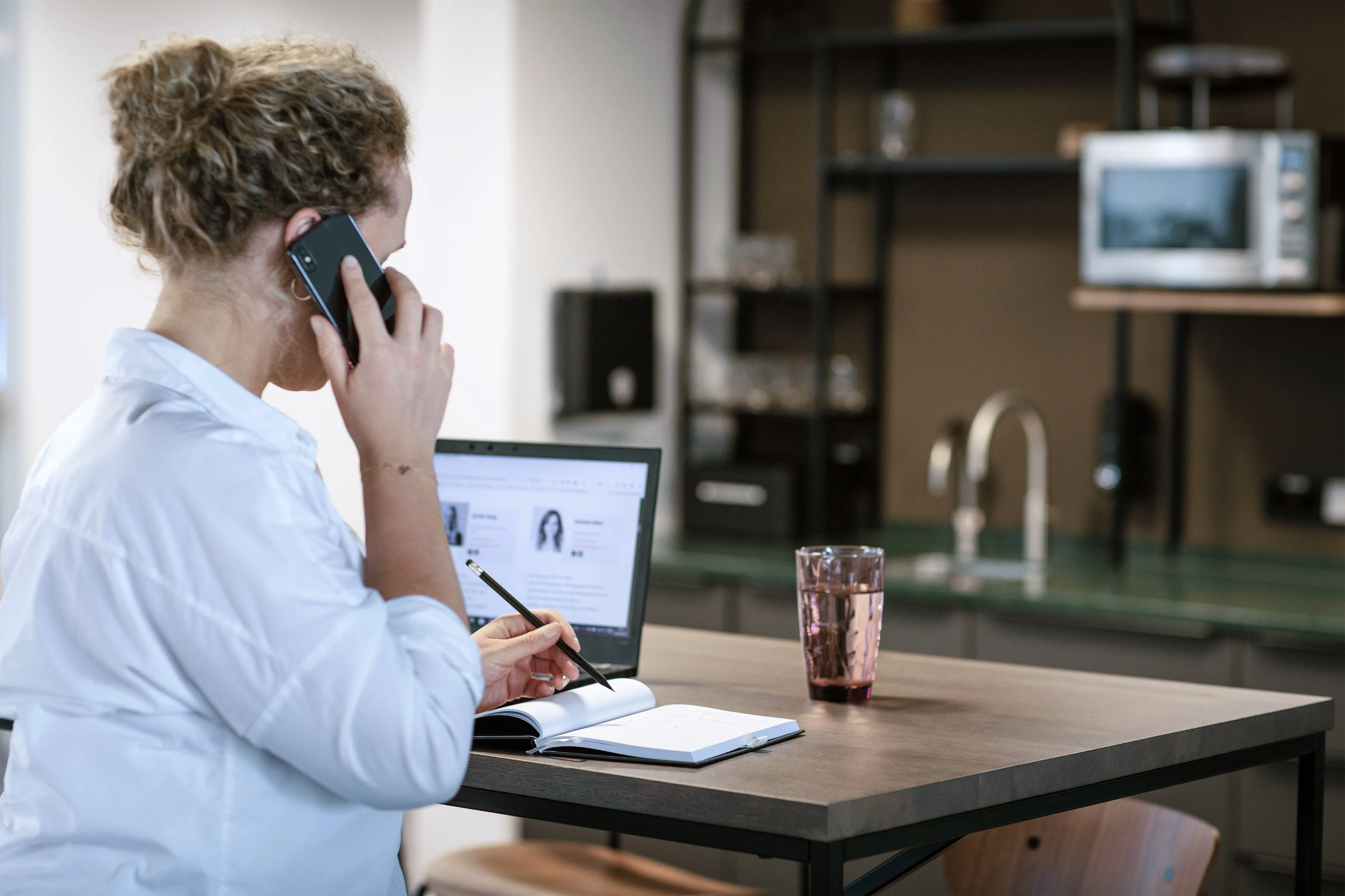 Woman in a casual setting multitasking with a phone call and taking notes on a laptop, capturing the dynamic work style at diva-e.