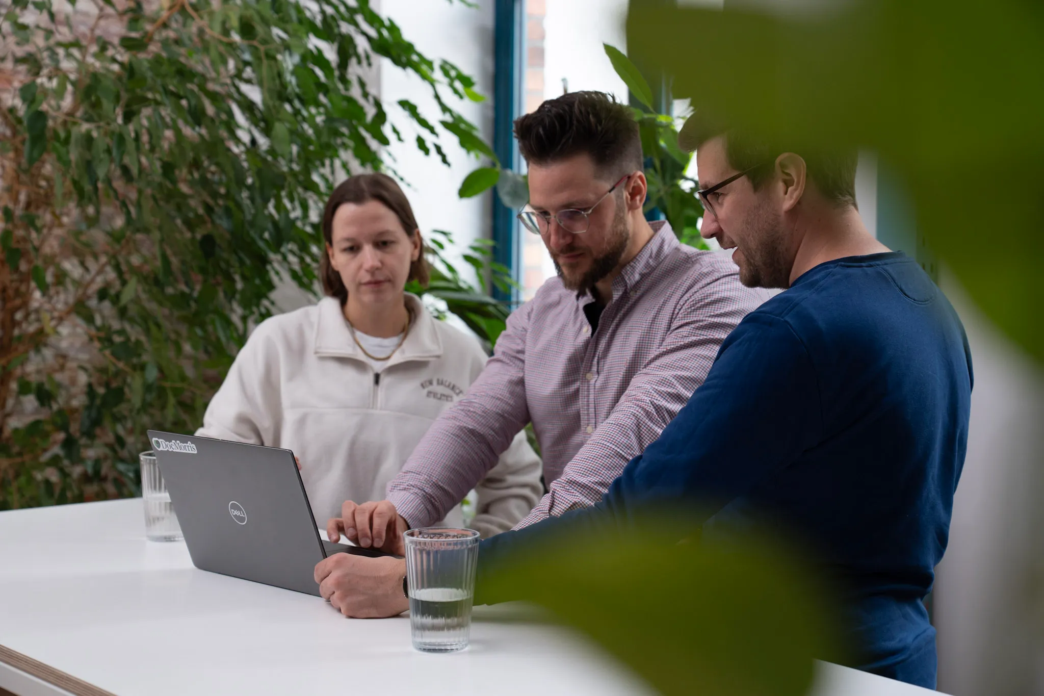 Drei Kollegen diskutieren an einem Laptop in einem modernen Büro mit Pflanzen im Hintergrund.
