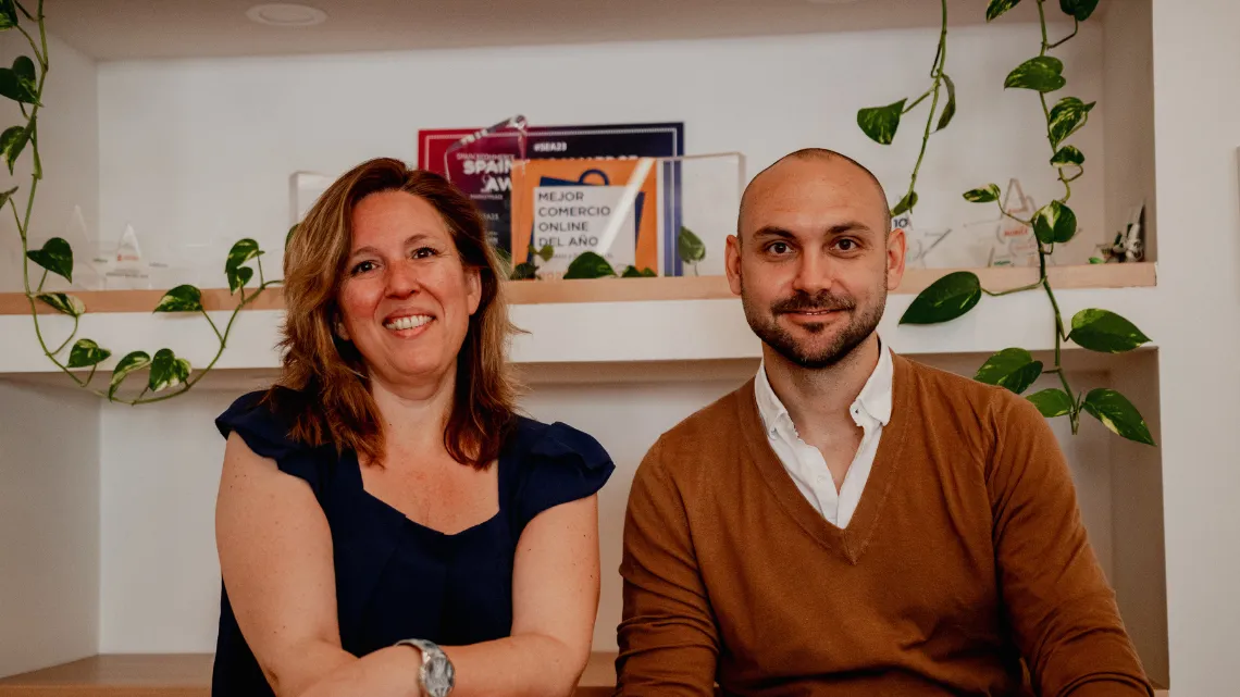 Dos personas sonríen en una oficina de PromoFarma by DocMorris, con premios de "Mejor Comercio Online" en el fondo, rodeados de plantas.