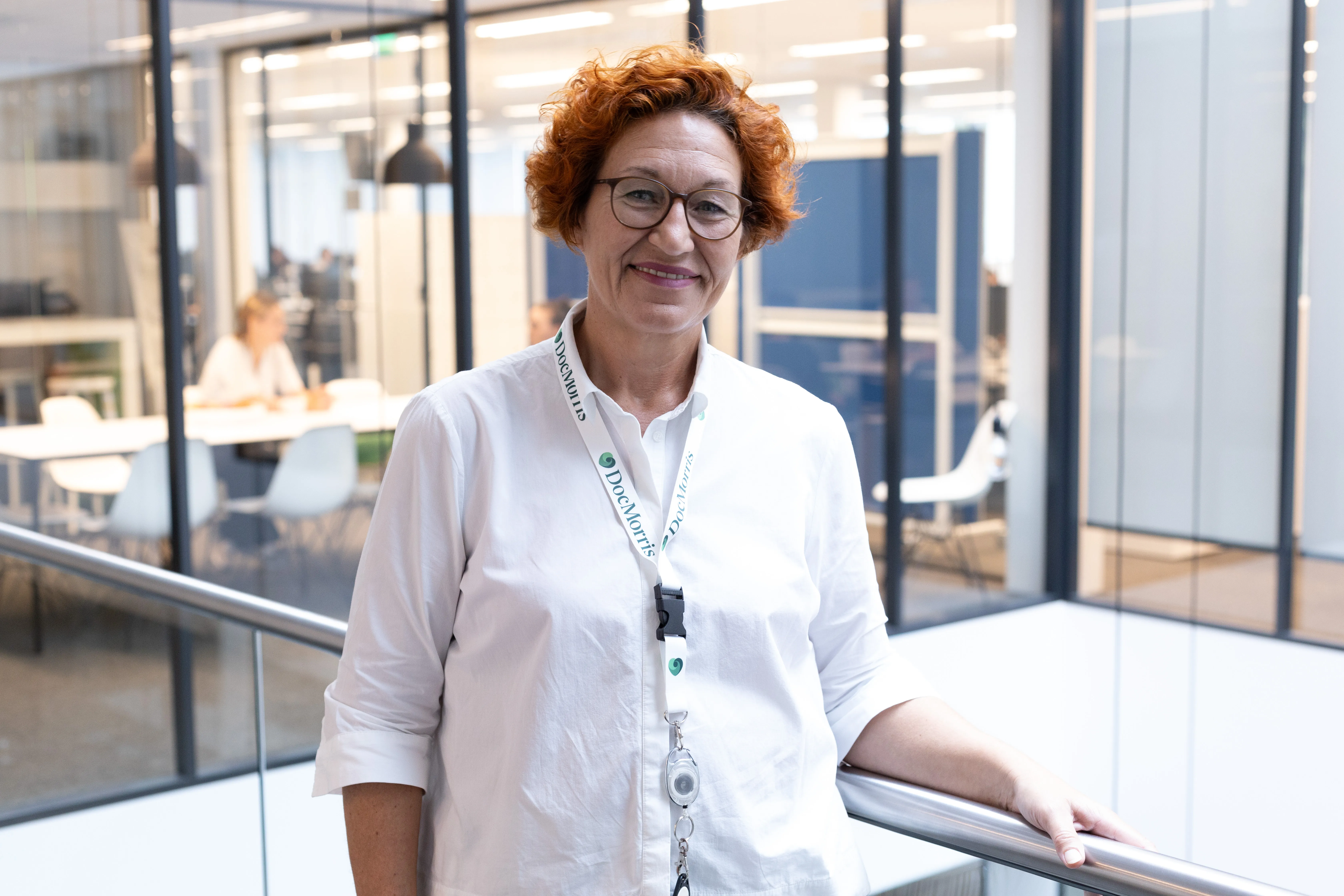 A friendly woman with curly hair smiles while leaning on a railing in a modern DocMorris office.