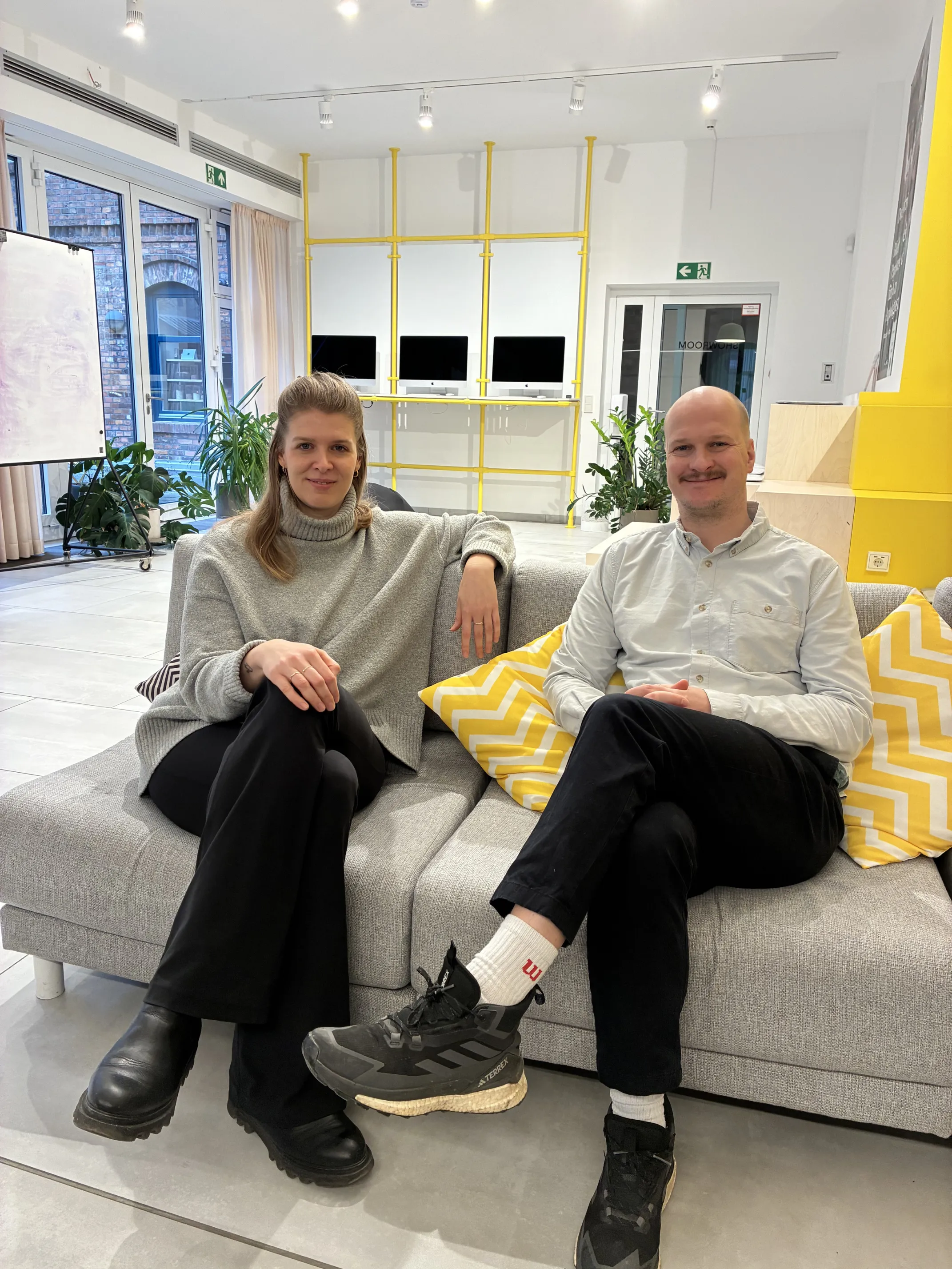 Zwei Personen sitzen entspannt auf einem Sofa in einem modernen Büro bei DocMorris, umgeben von Pflanzen und Computern.