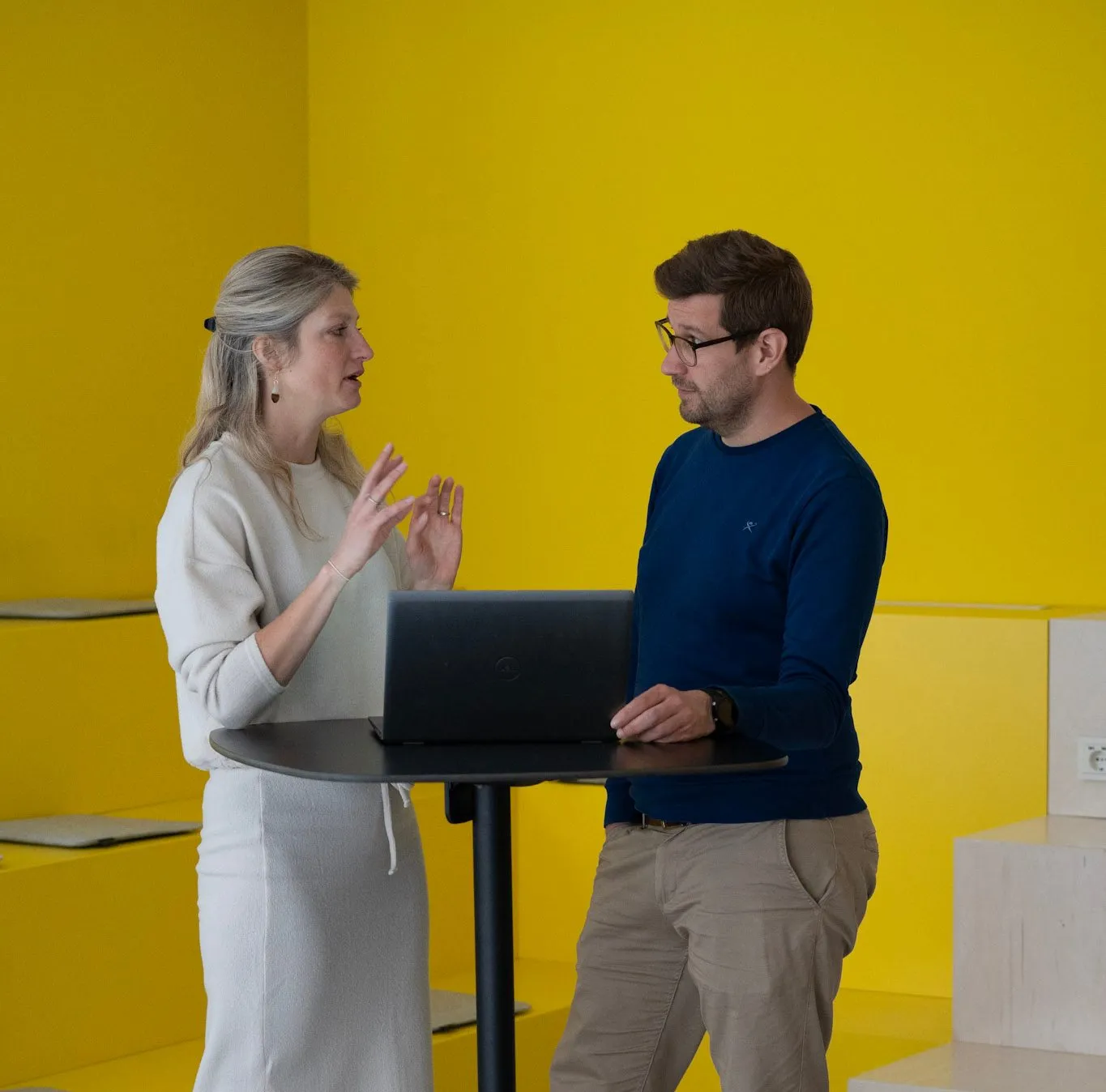 Zwei Personen diskutieren an einem Stehtisch mit Laptop vor einer gelben Wand, in einem Büro von DocMorris.