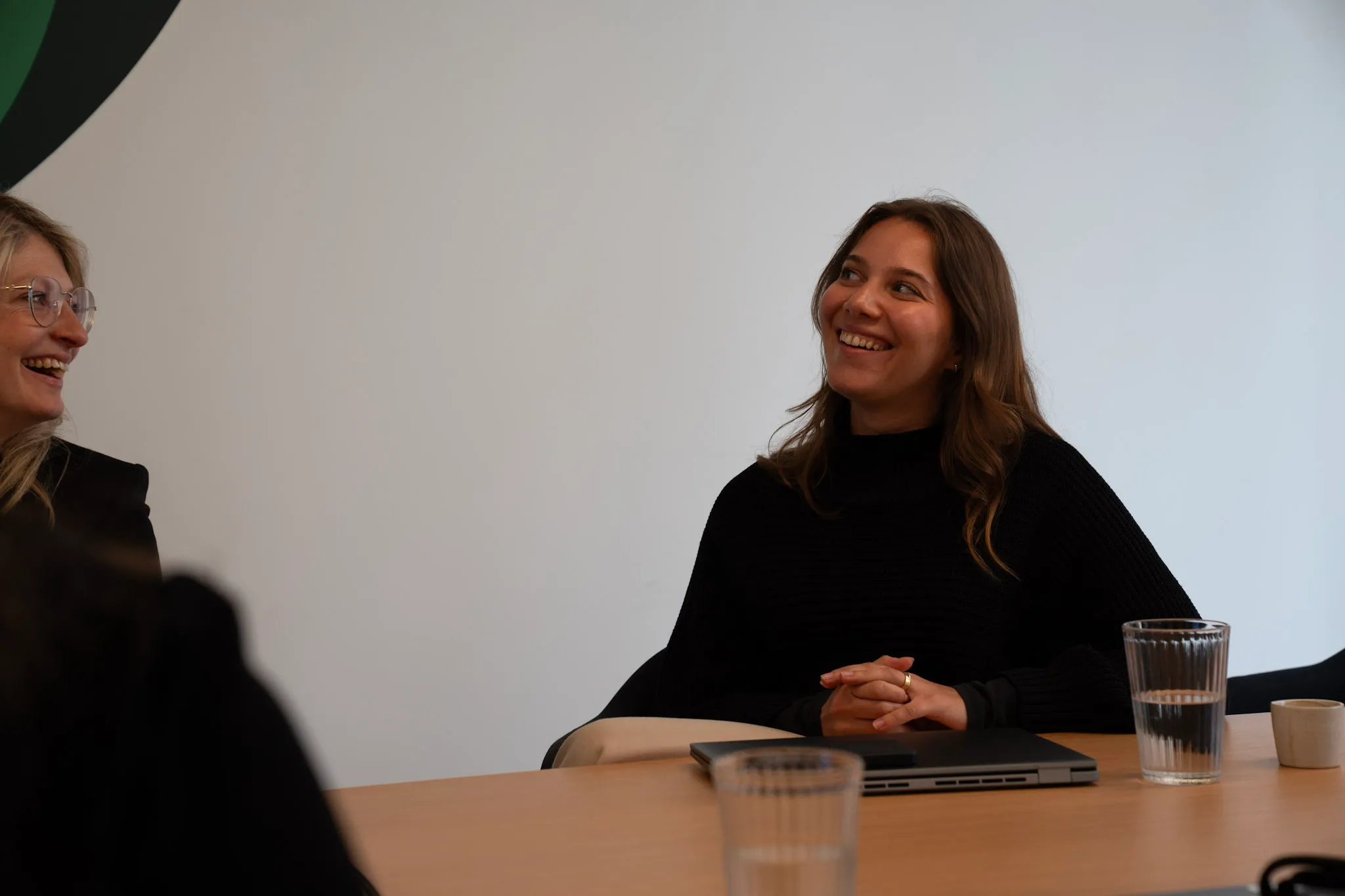 Zwei lächelnde Frauen in einem Meeting bei DocMorris, mit Laptop und Wassergläsern auf dem Tisch.