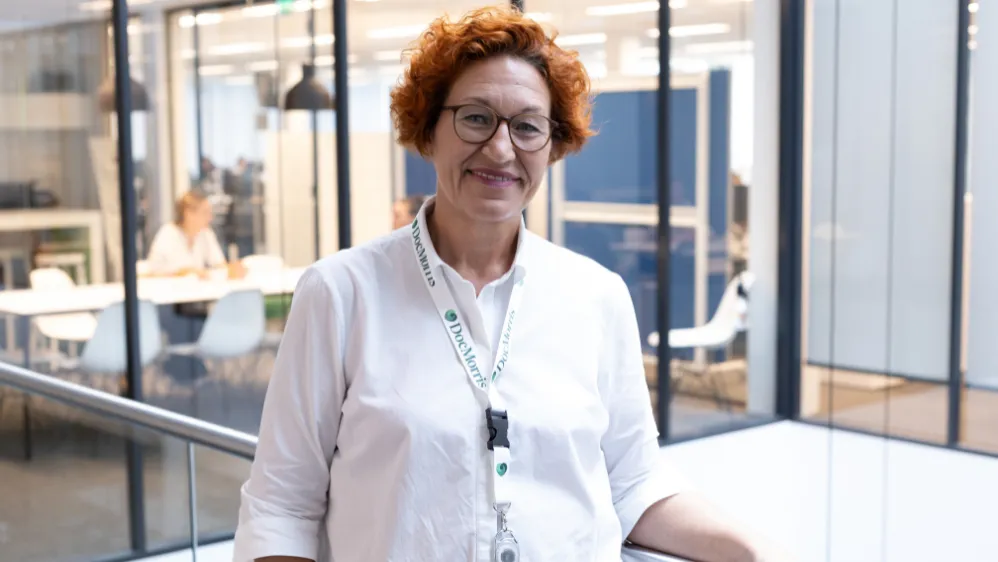 A friendly DocMorris employee with curly hair and glasses smiles in a modern office setting.