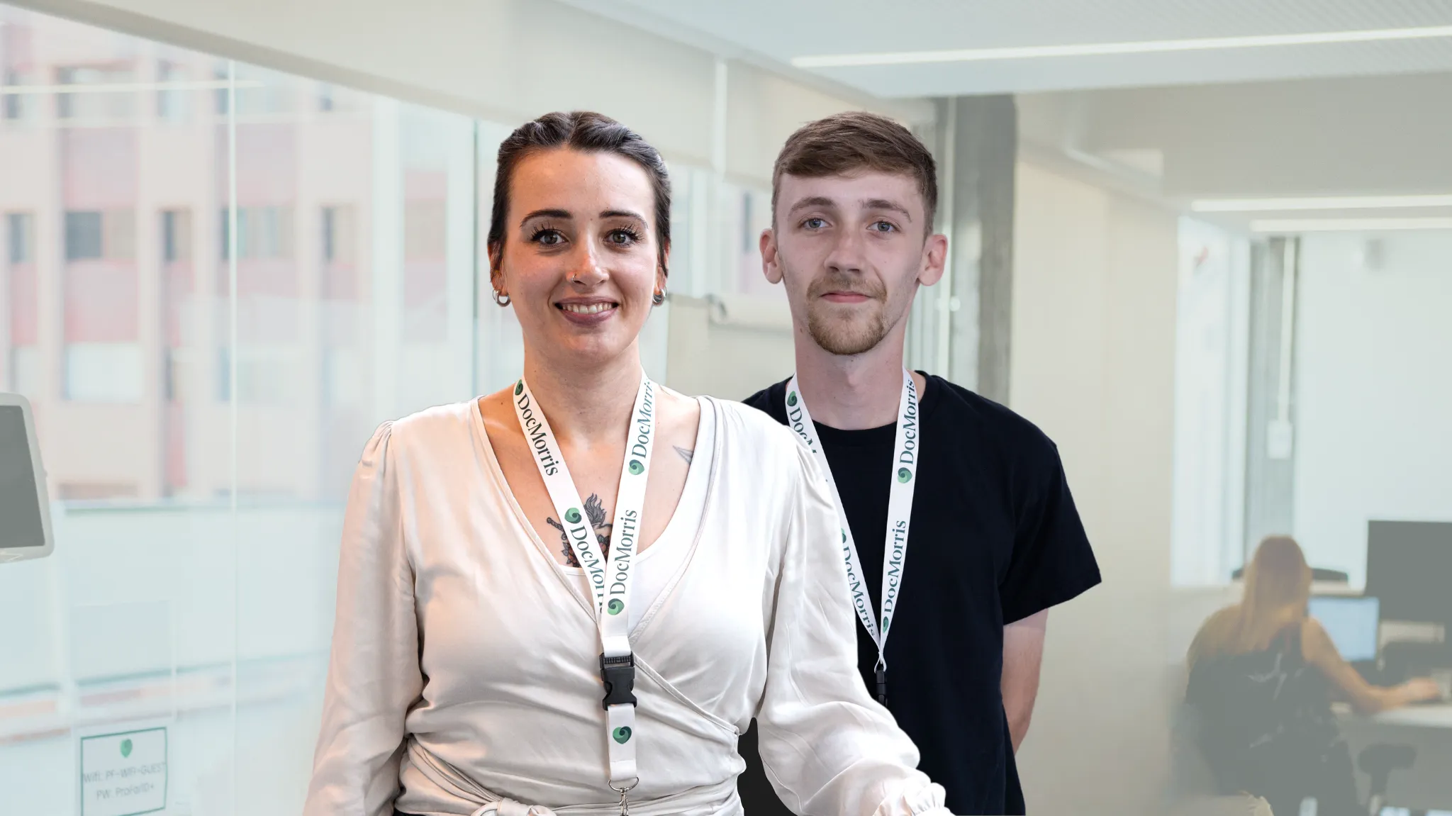 Two friendly DocMorris employees, wearing branded lanyards, smiling in a modern office setting.