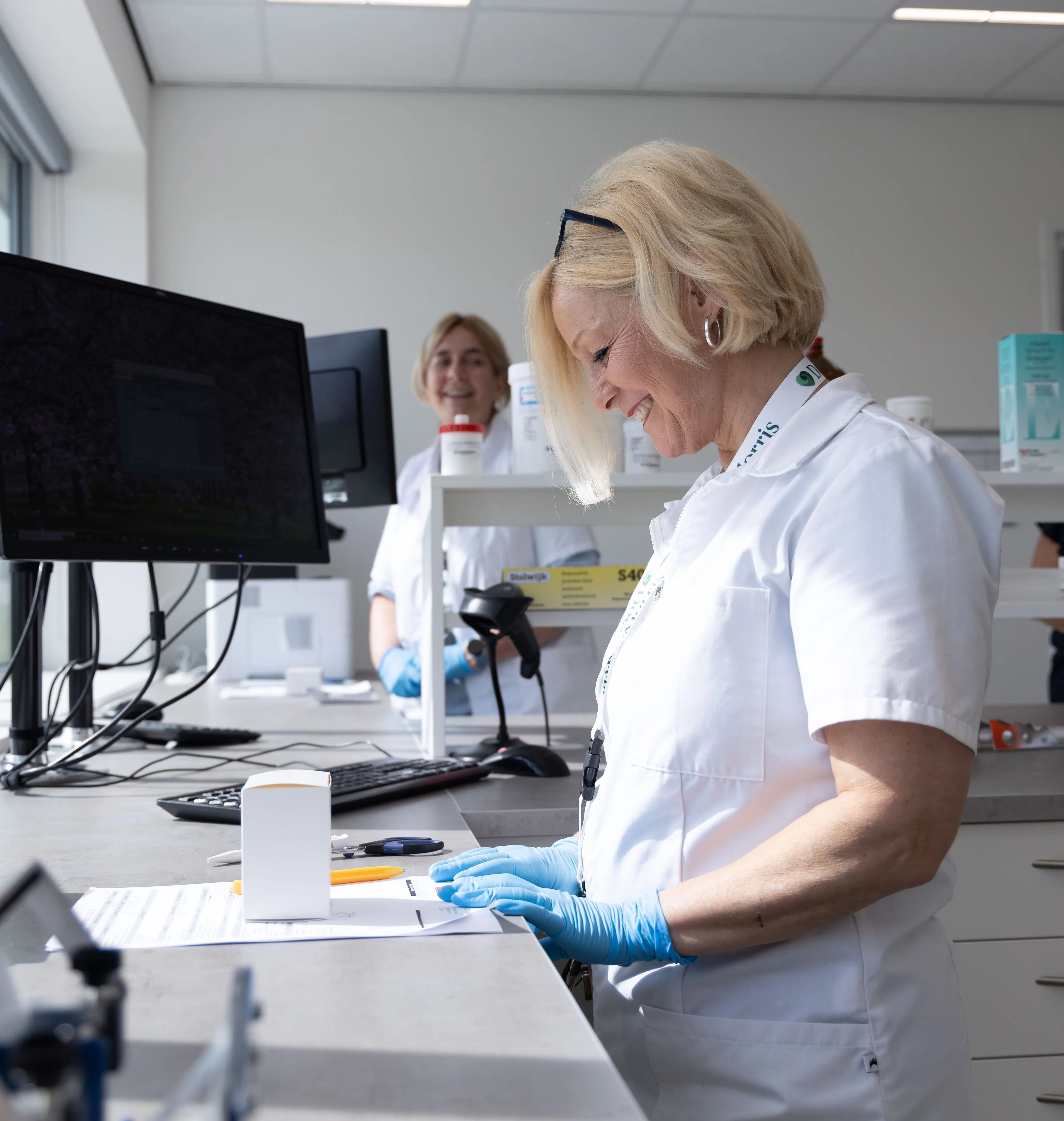 Two cheerful professionals in a pharmacy setting, wearing lab coats and gloves, focus on their tasks at DocMorris.