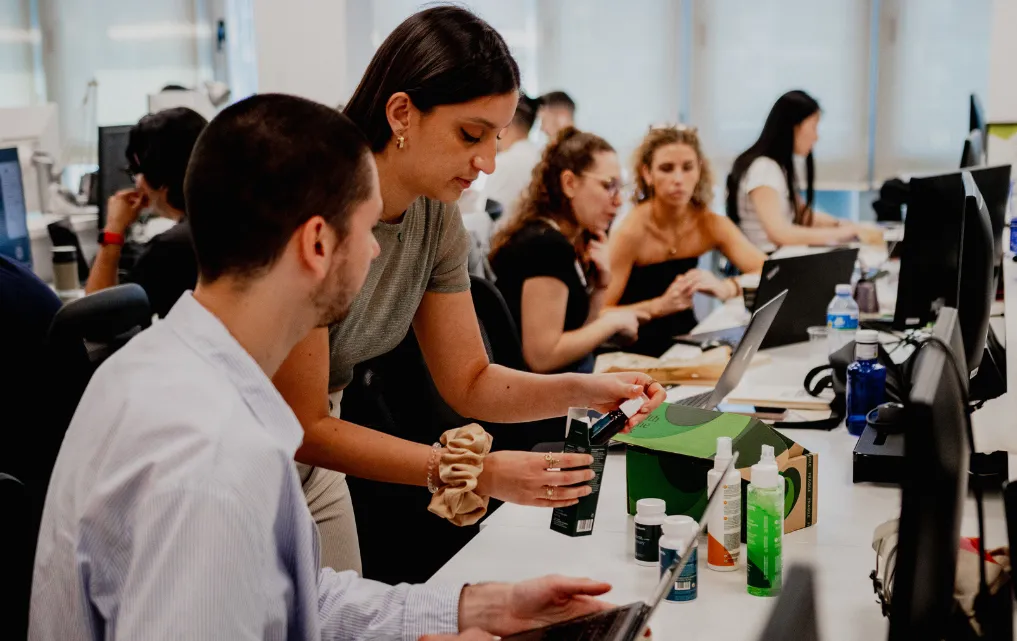 Un grupo de jóvenes profesionales en una oficina de PromoFarma revisa productos farmacéuticos en sus escritorios.