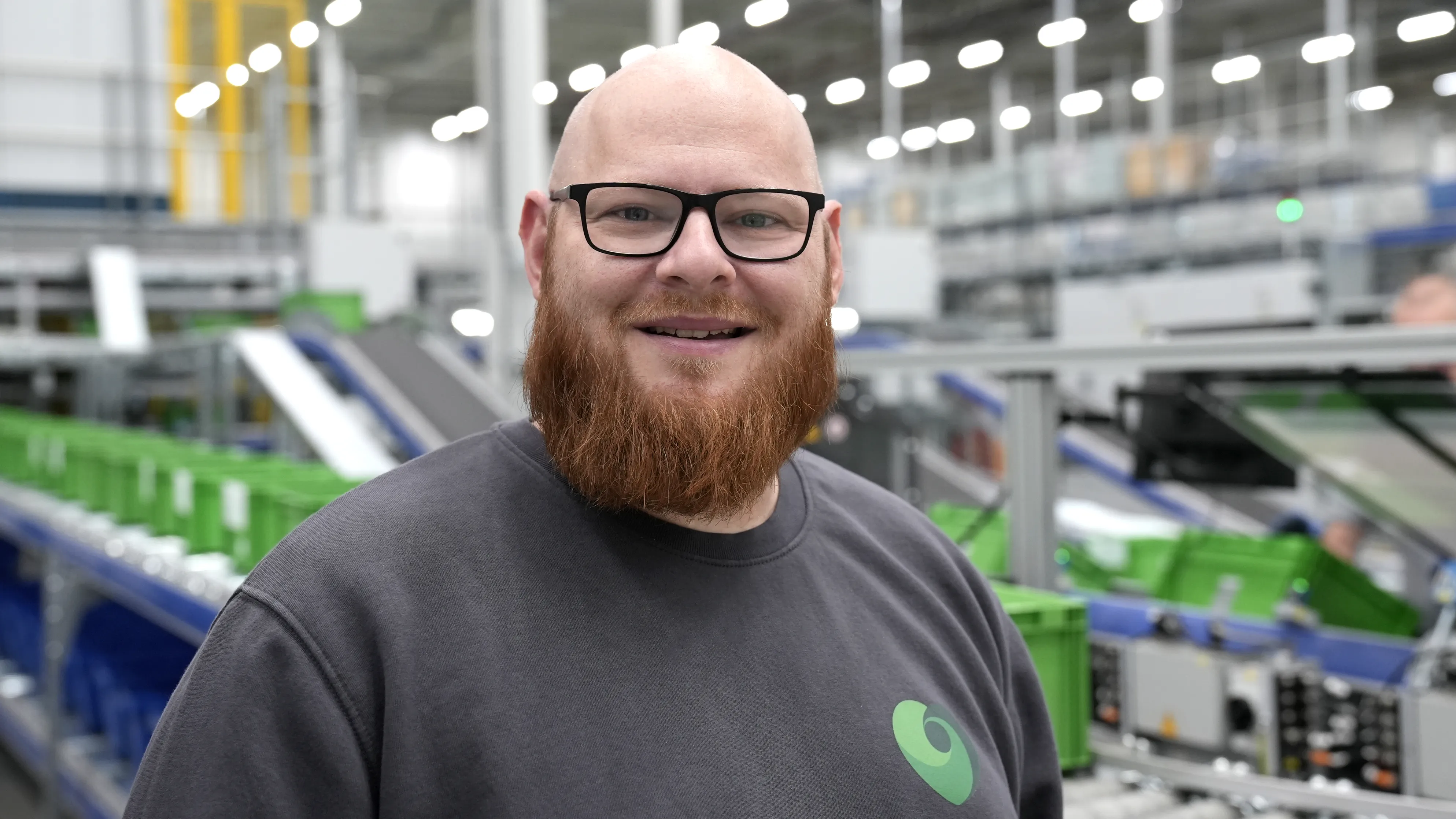 A friendly DocMorris employee with curly hair and glasses smiles in a modern office setting.