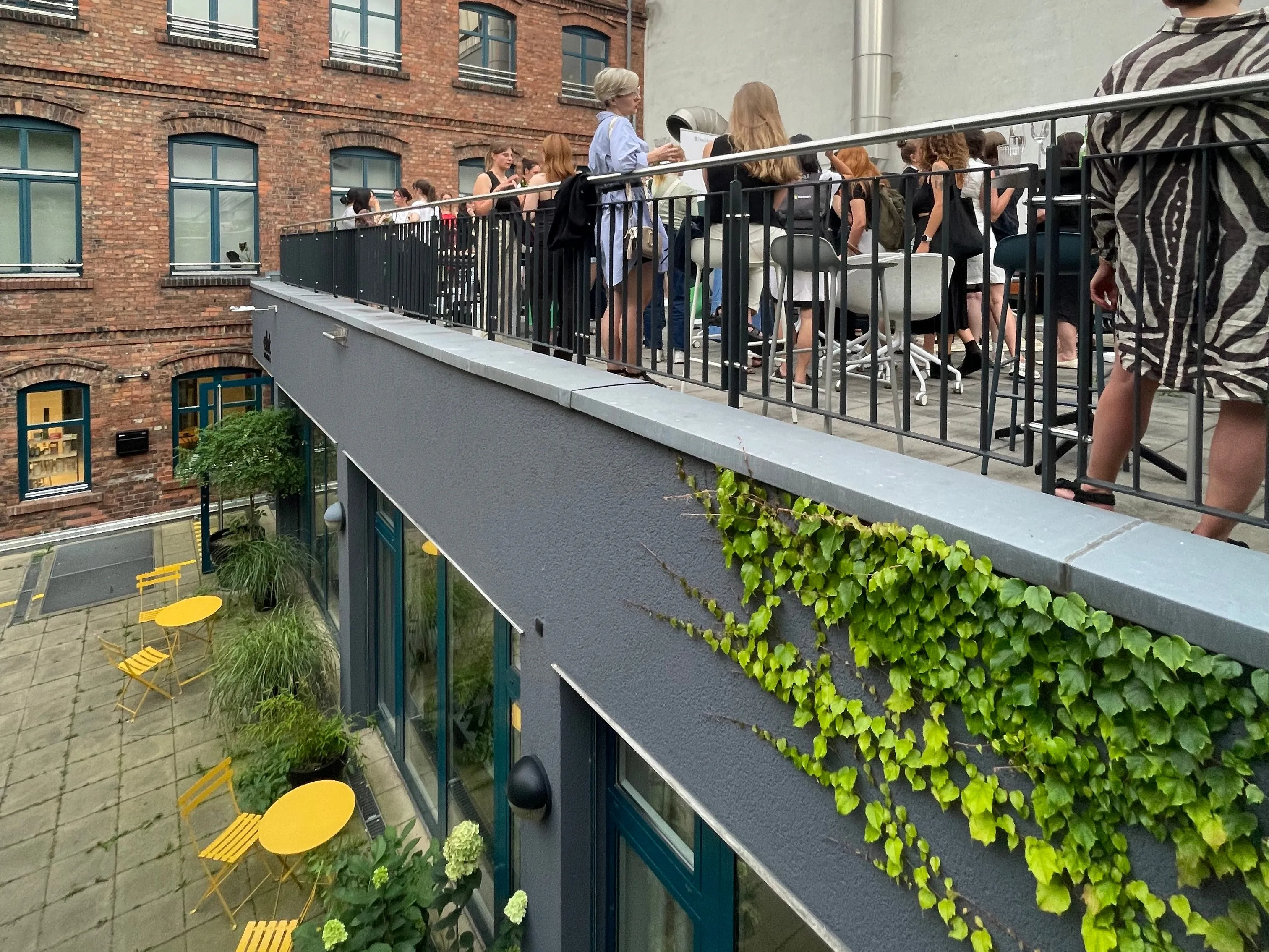 Outdoor gathering on a modern terrace with people networking, surrounded by greenery and vibrant yellow chairs below.