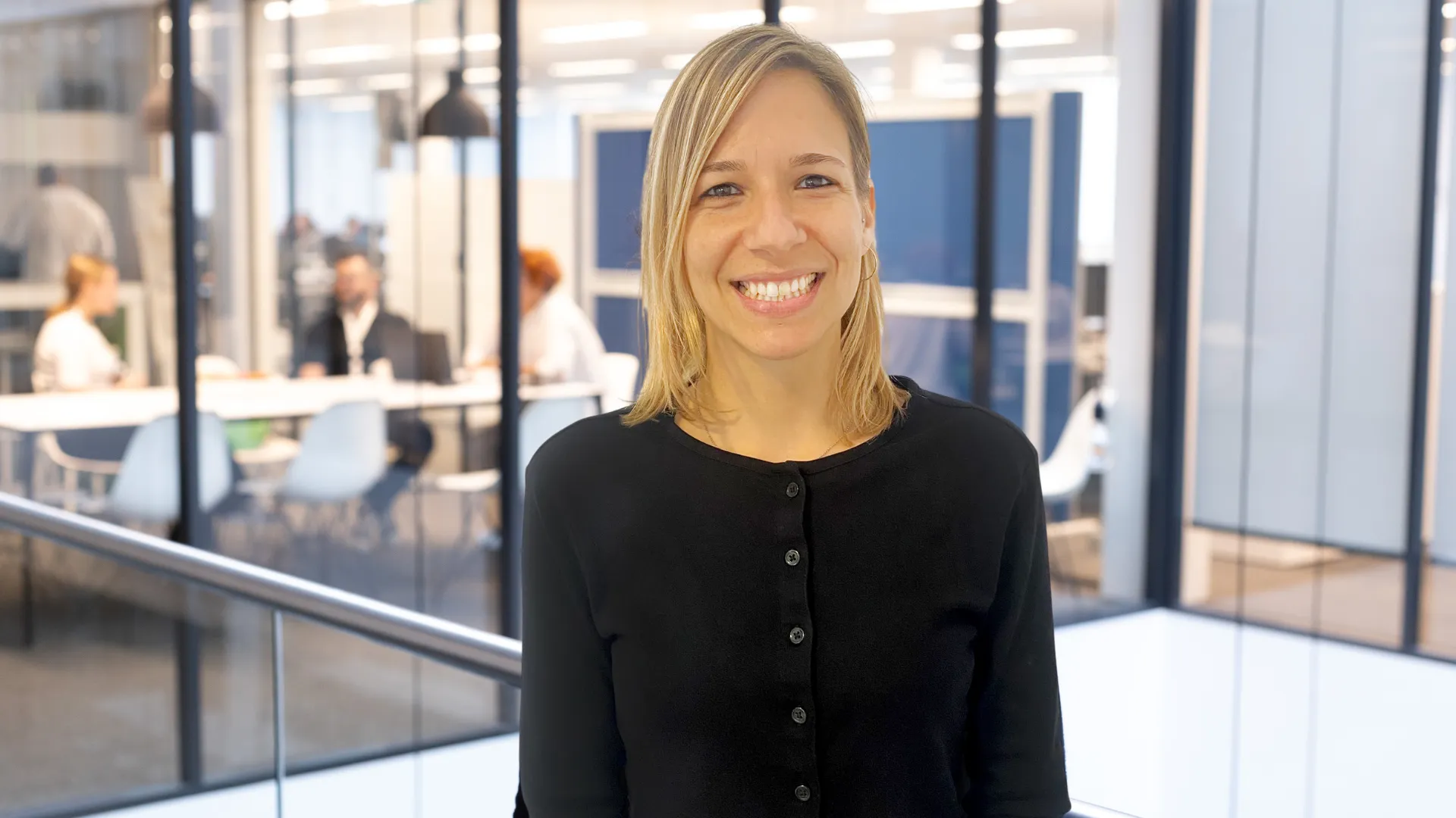 A smiling professional in a black top stands in a modern office with colleagues in the background.