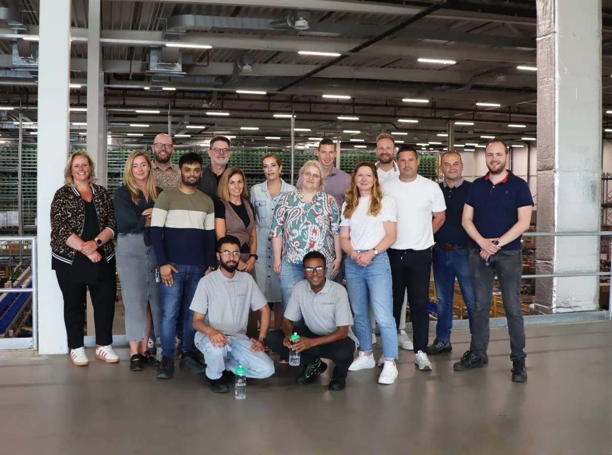 A smiling professional in a black top stands in a modern office with colleagues in the background.