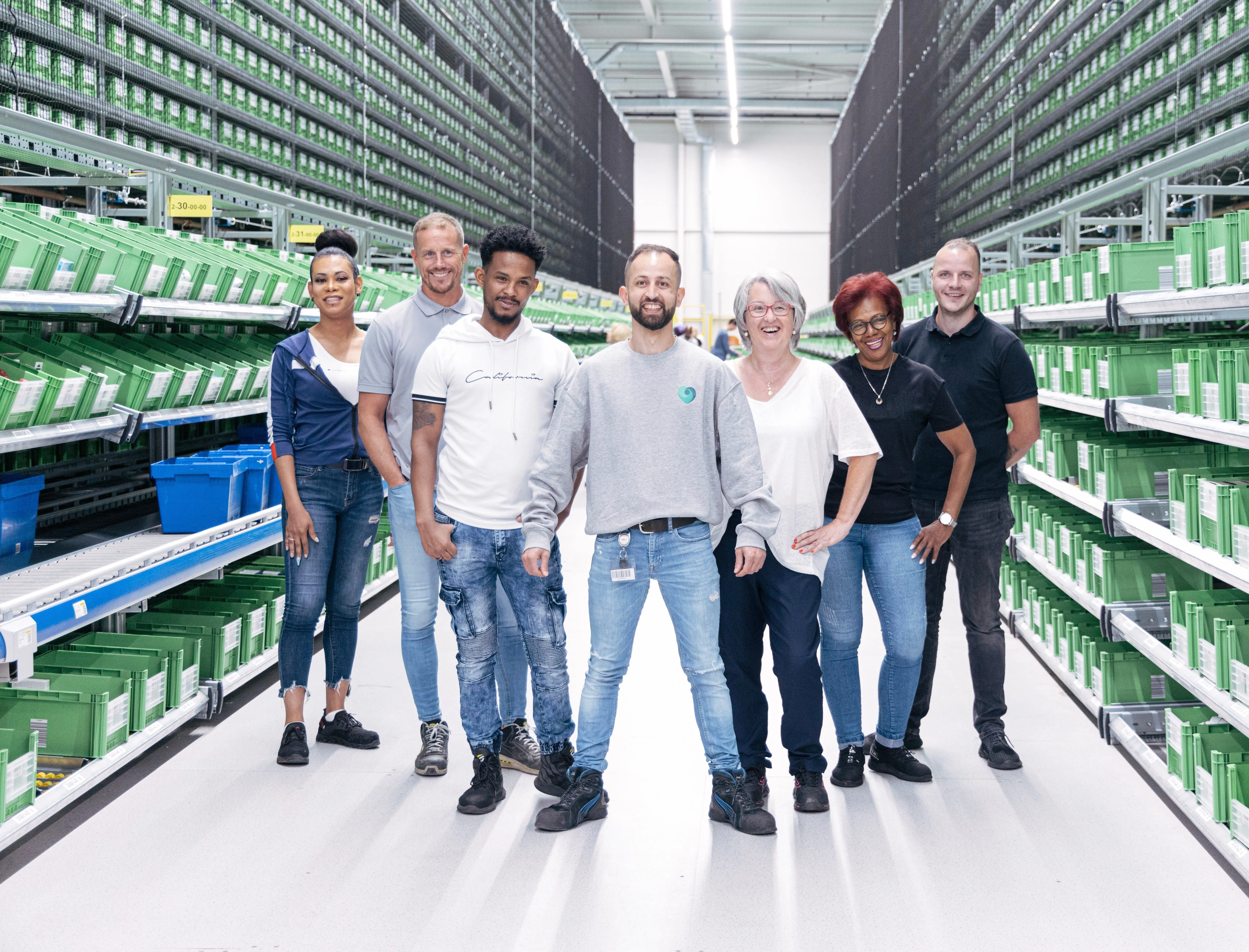 A friendly team of DocMorris employees standing in a warehouse aisle, surrounded by shelves filled with green bins.