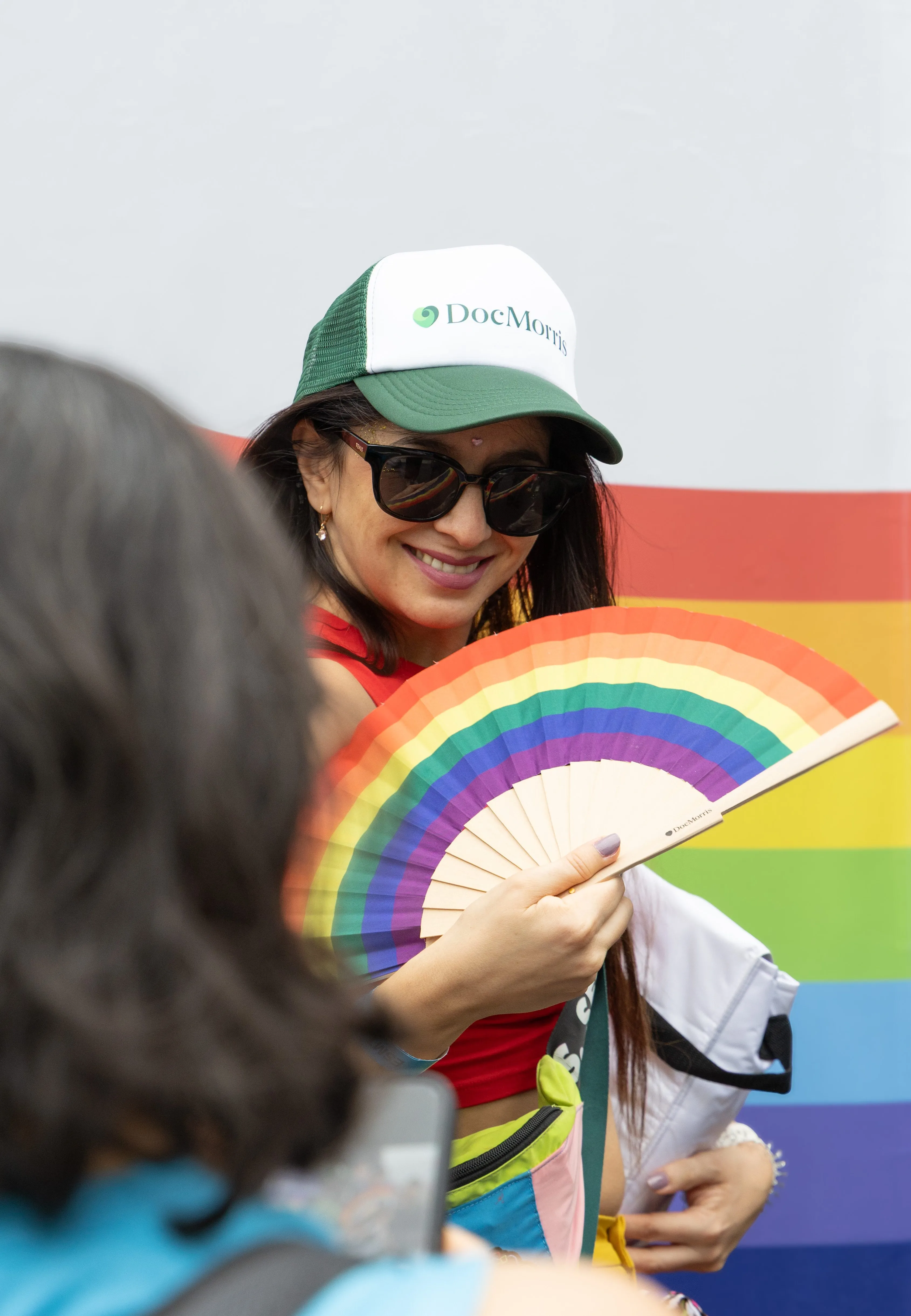 Woman in a DocMorris cap smiling and holding a rainbow fan, enjoying a healthcare event.