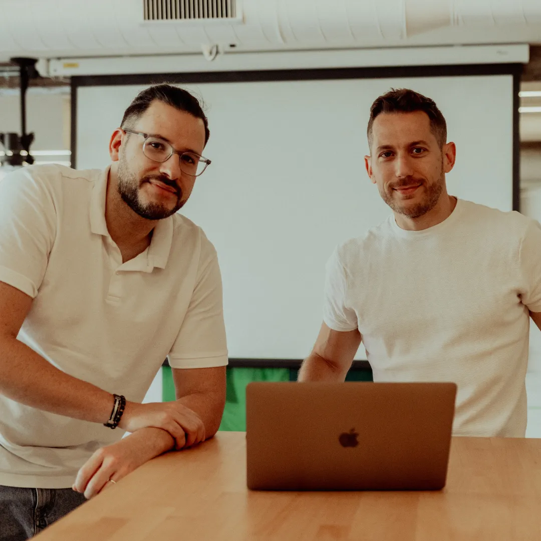 Two friendly guys in casual white shirts, chilling at a wooden table with a laptop, ready to collaborate in healthcare at DocMorris.