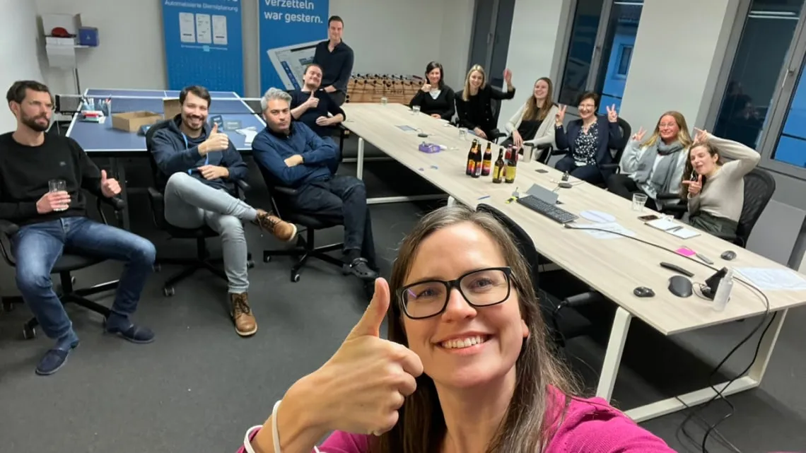 Fröhliches Team der doctari group im Büro, locker sitzend um einen Tisch, gibt Daumen hoch und lacht in die Kamera.