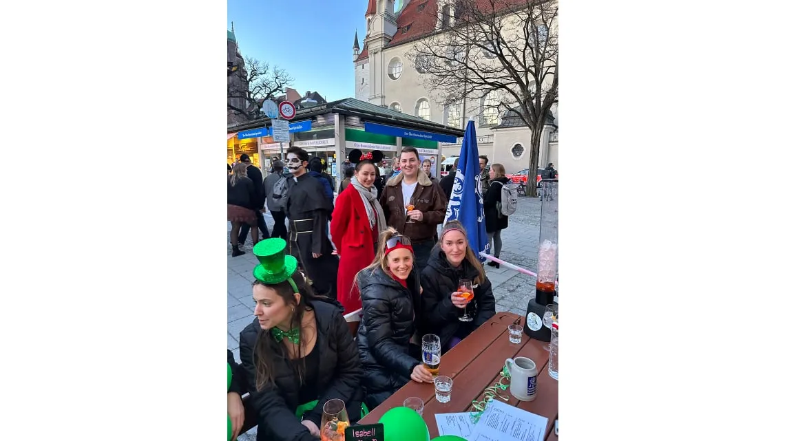 Mehrere junge Leute feiern kostümiert draußen an einem Tisch, lachen und stoßen mit Getränken an.
