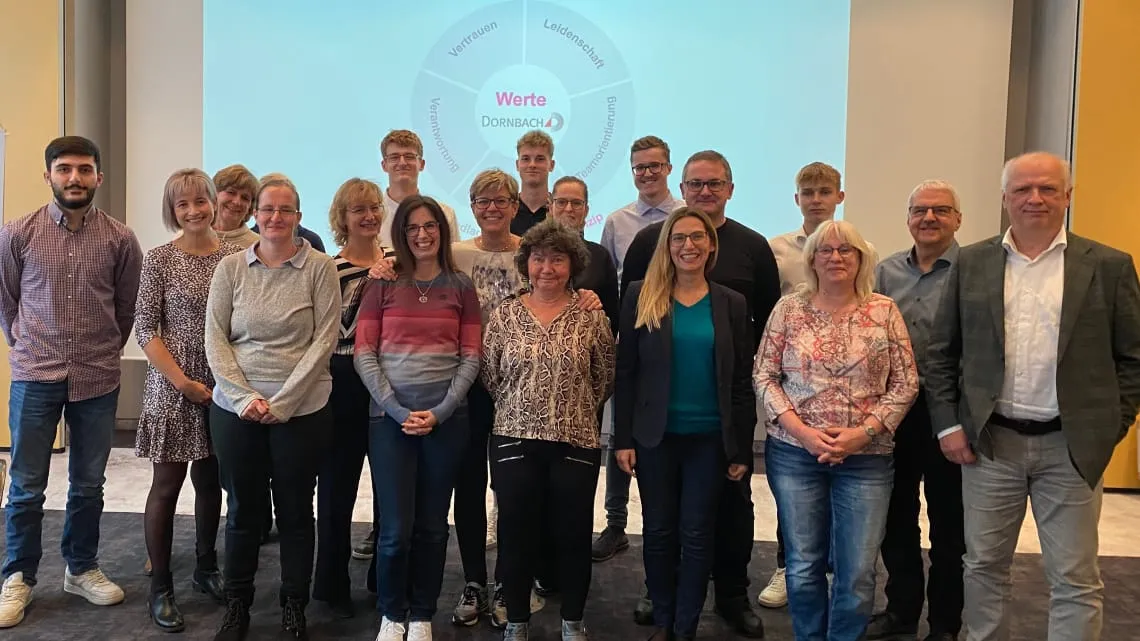Eine Gruppe von DORNBACH Mitarbeitenden lächelt gemeinsam für ein Teamfoto im Büro vor einer Präsentationsfolie mit Unternehmenswerten.