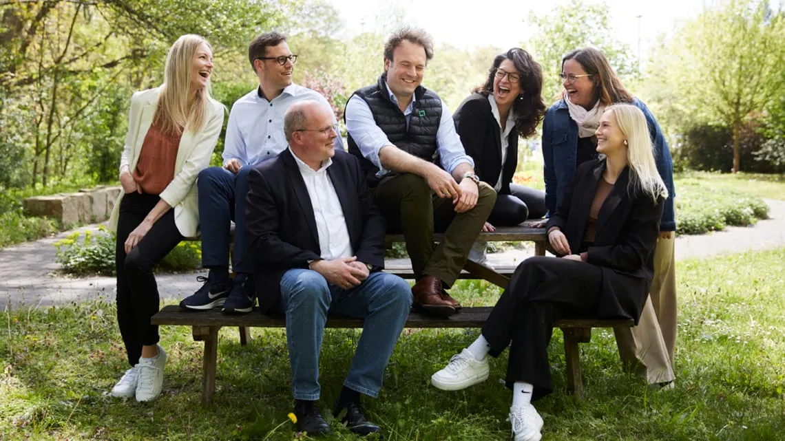 Sieben Kolleg*innen von ganz meine Natur sitzen lachend draußen zusammen und zeigen Teamspirit in der Pharma-Branche.