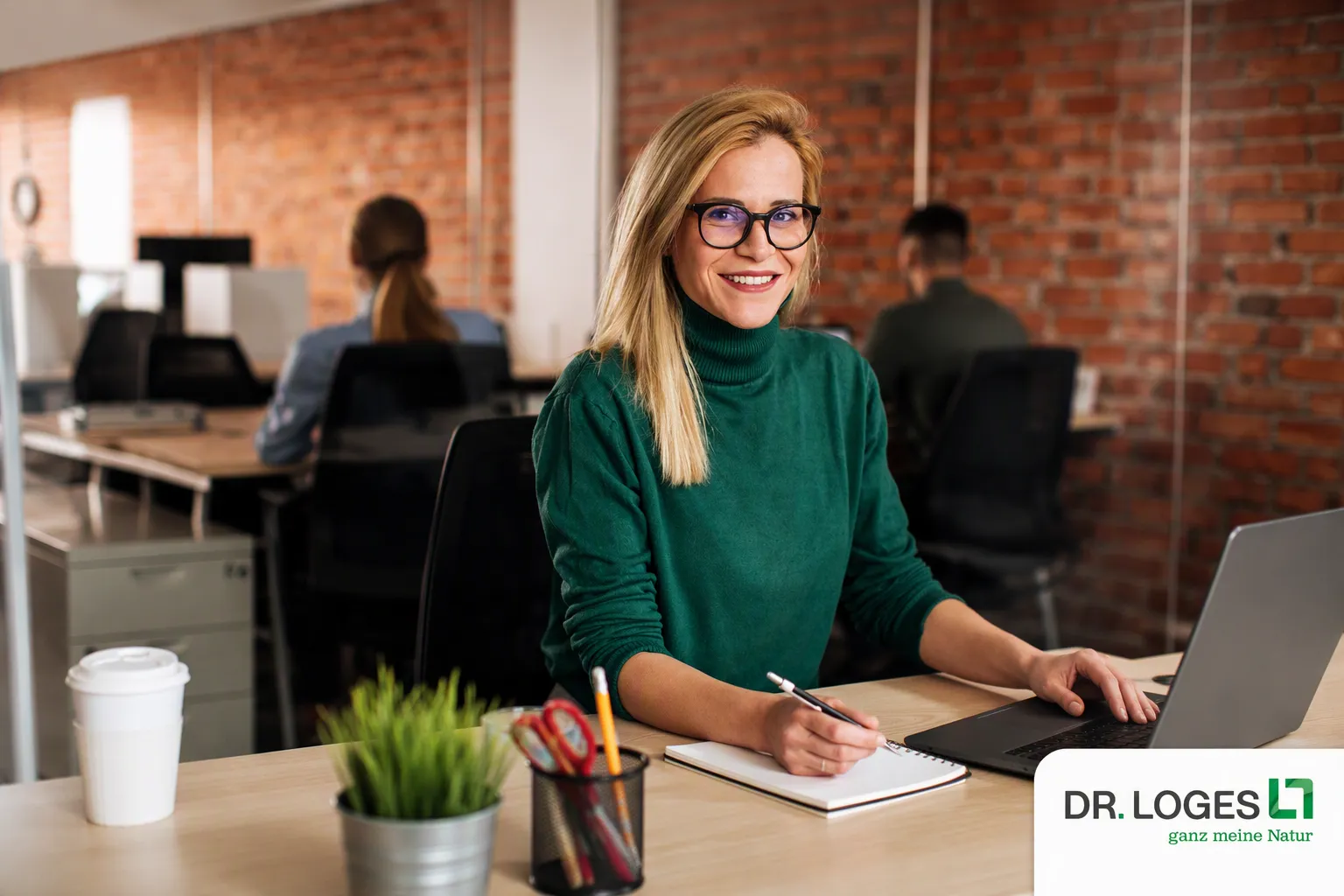 Junge Frau mit Brille arbeitet lächelnd am Schreibtisch im modernen Büro bei Dr. Loges – entdecke Deine Karriere in der Pharmaindustrie.