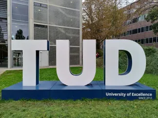 Große TUD-Buchstaben auf dem Campus der Technischen Universität Dresden, umgeben von grüner Wiese und Gebäuden.