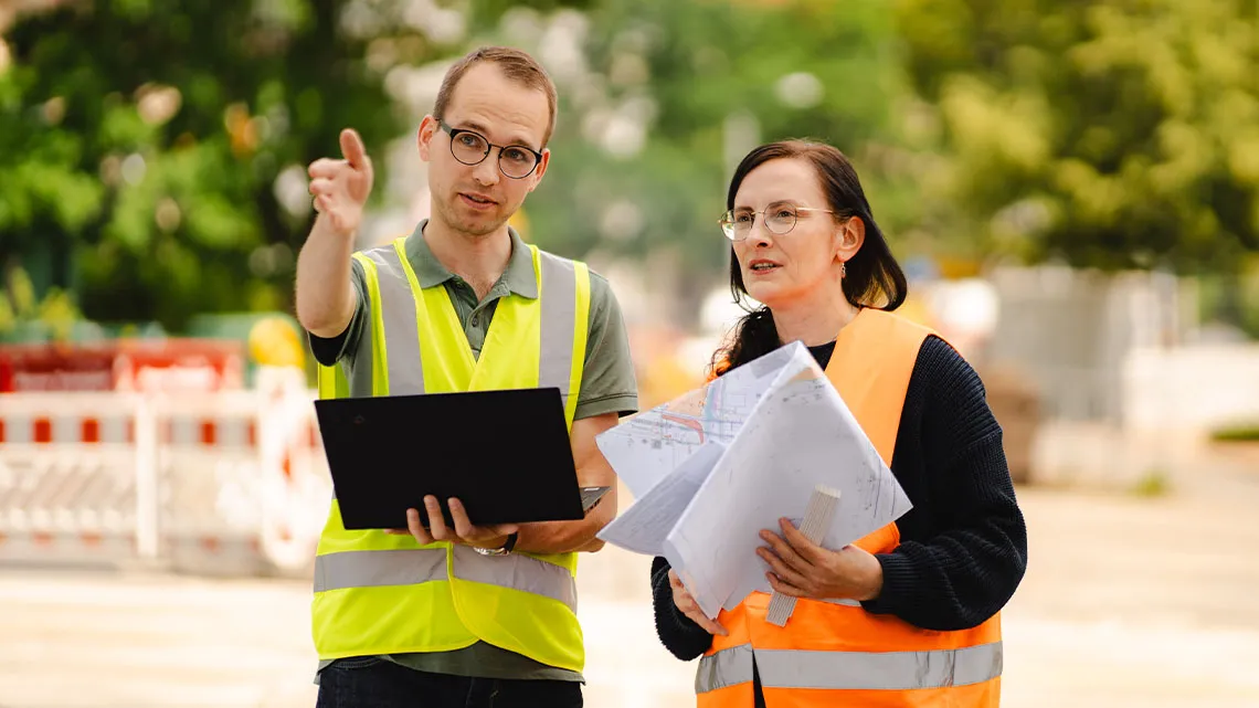 Zwei Personen in Warnwesten besprechen Baupläne im Freien, einer zeigt mit der Hand, während Du zuhörst.