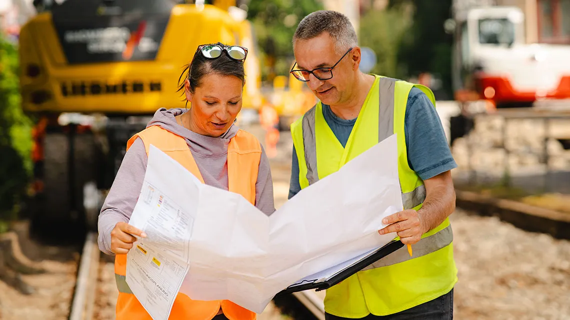 Zwei Bauleiter besprechen gemeinsam Pläne auf einer Baustelle, Du siehst Zusammenarbeit im Arbeitsalltag.