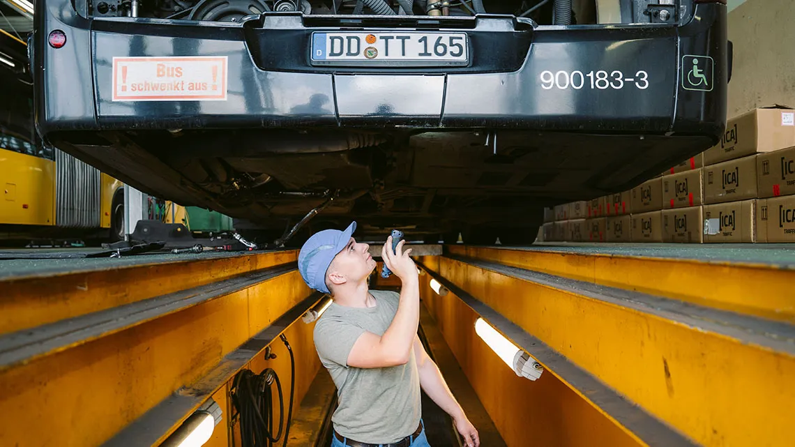 Du siehst einen Mechaniker, der mit einer Taschenlampe unter einem angehobenen Bus in einer Werkstatt arbeitet.