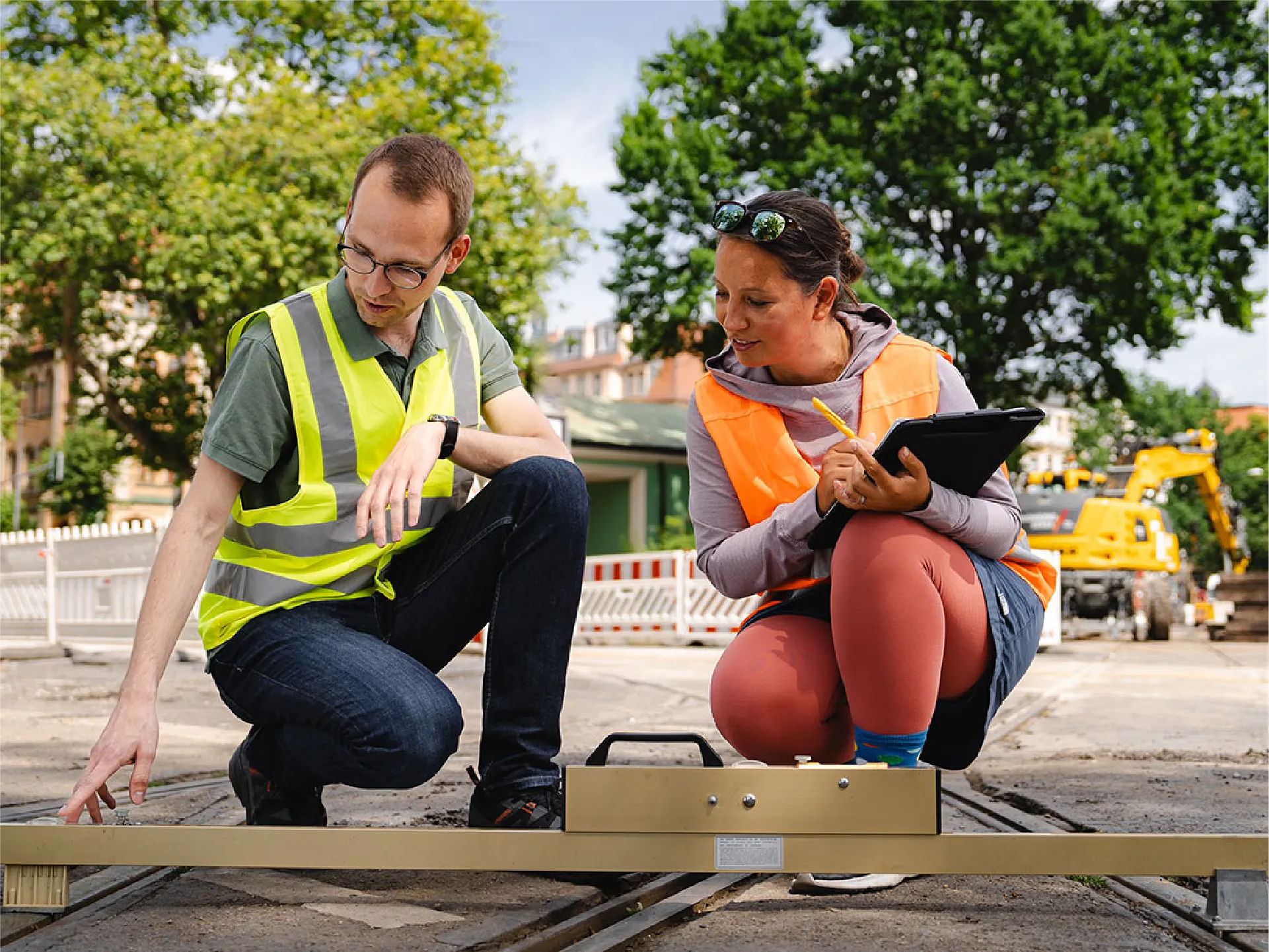 Zwei Ingenieure in Warnwesten arbeiten im Freien an einer Baustelle. Einer zeigt auf die Schienen, während die andere Notizen auf einem Tablet macht.