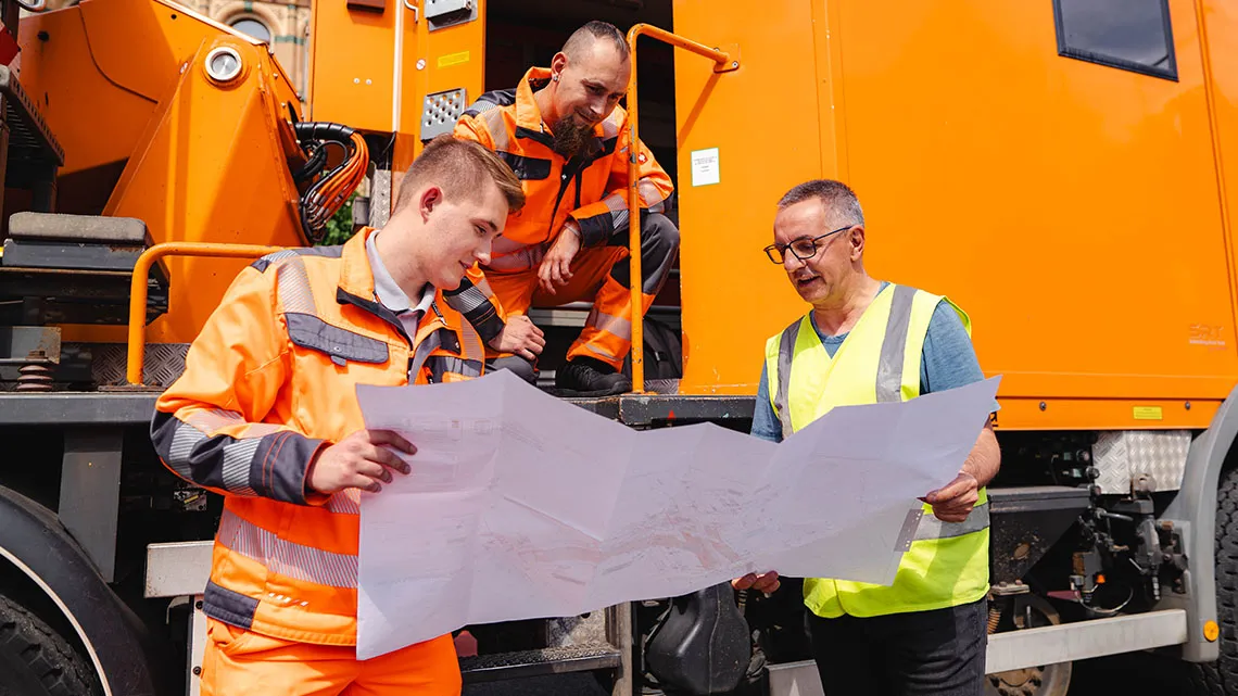 Drei Arbeiter in Warnkleidung besprechen gemeinsam Baupläne vor einem orangefarbenen Einsatzfahrzeug.