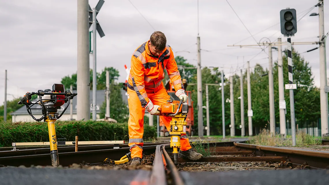 Du siehst einen Arbeiter in orangefarbener Schutzkleidung, der Gleisarbeiten mit einer Maschine durchführt.