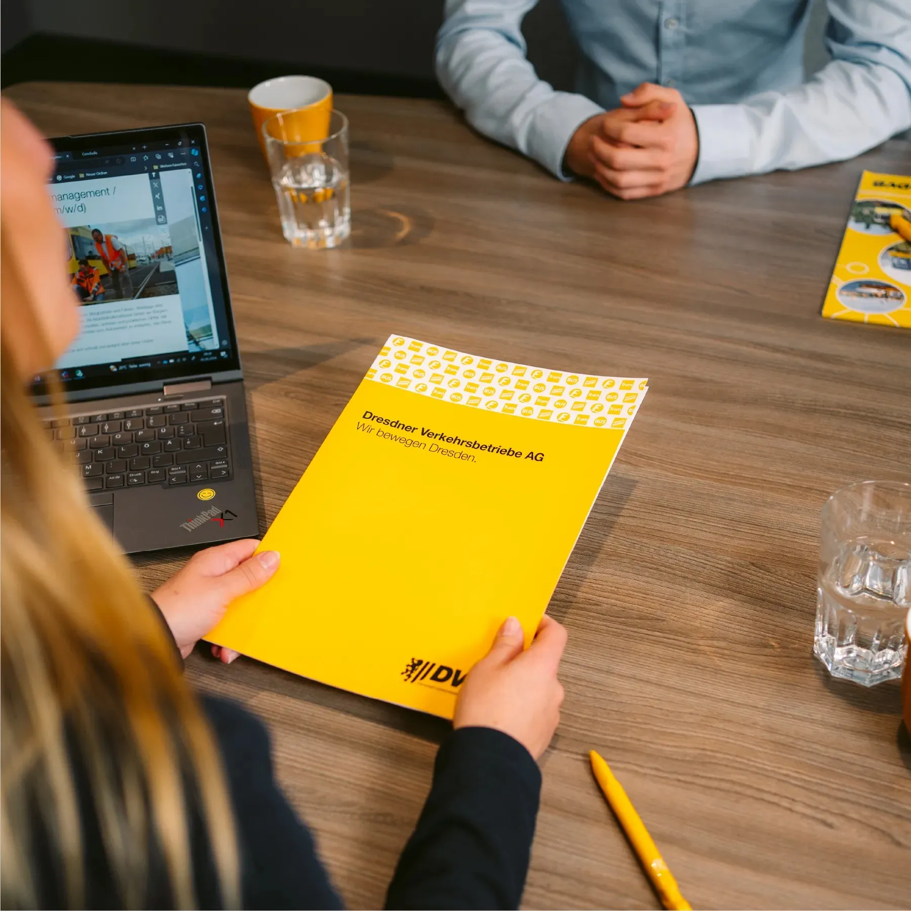 A person holds a yellow brochure from Dresdner Verkehrsbetriebe AG at the conference table, next to a laptop and glasses of water.
