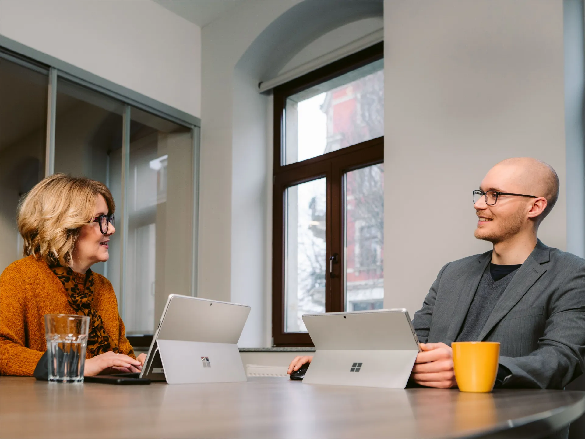 Zwei Personen in einem Büro sitzen an einem Tisch und arbeiten mit Laptops. Sie lächeln und unterhalten sich.