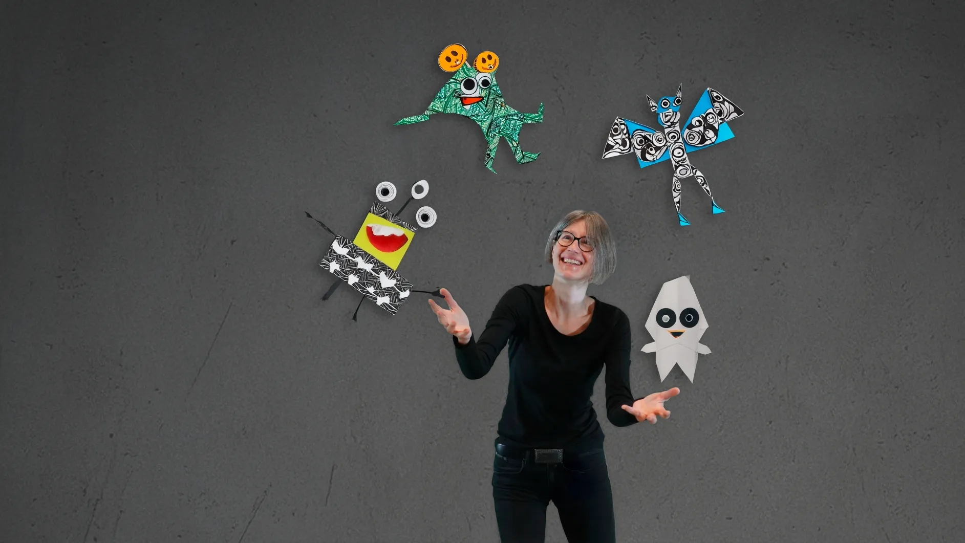 Eine lächelnde Frau jongliert bunte, gebastelte Monsterfiguren vor grauem Hintergrund.