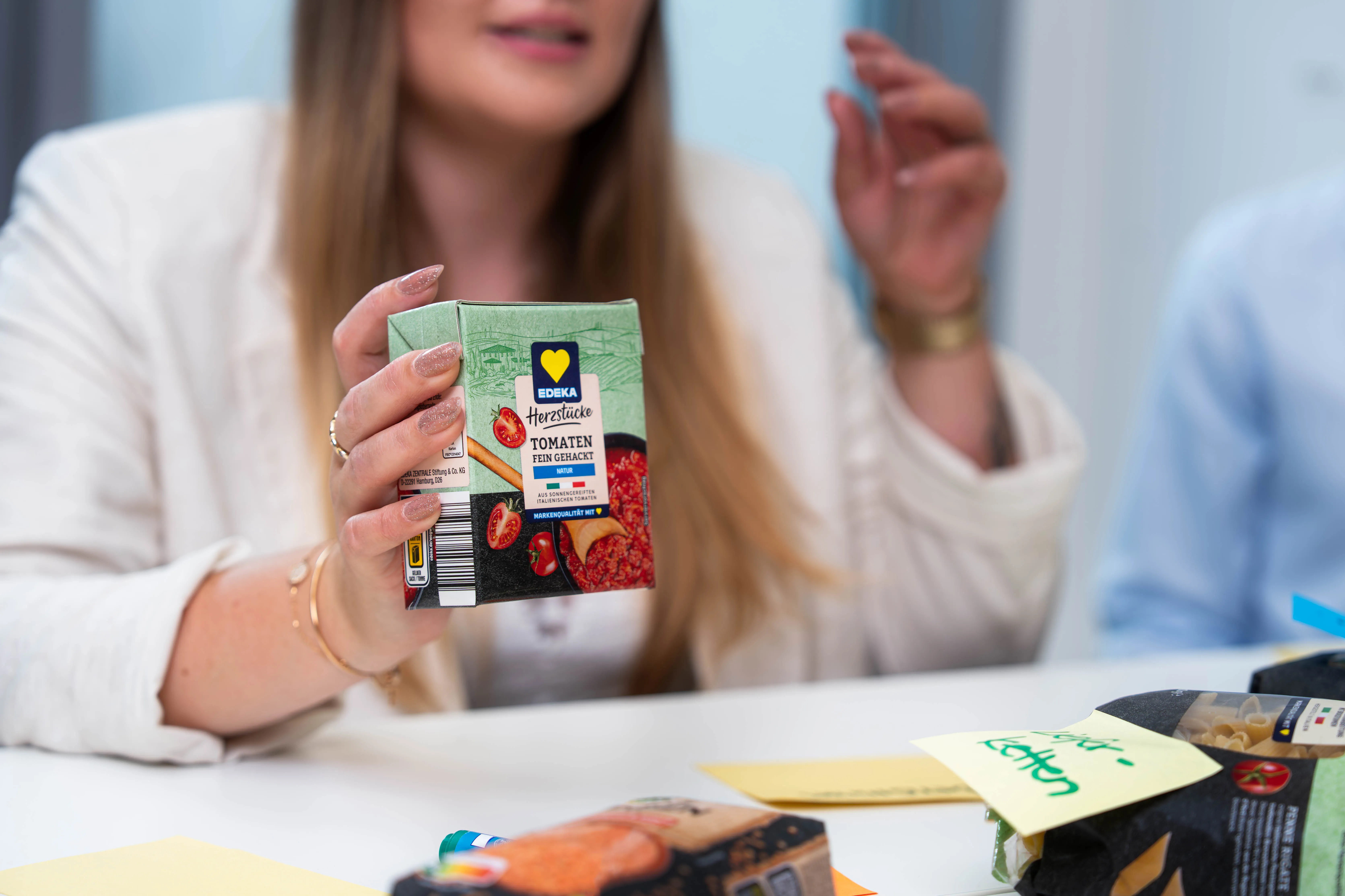 Eine lächelnde Frau sitzt in einem Büro und hält eine Tasse, während im Hintergrund Kollegen arbeiten.