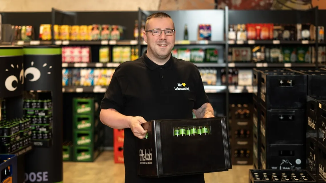 Mitarbeiter im EDEKA-Markt trägt Getränkekiste und lächelt, du siehst ihn vor den Regalen im Getränkebereich.