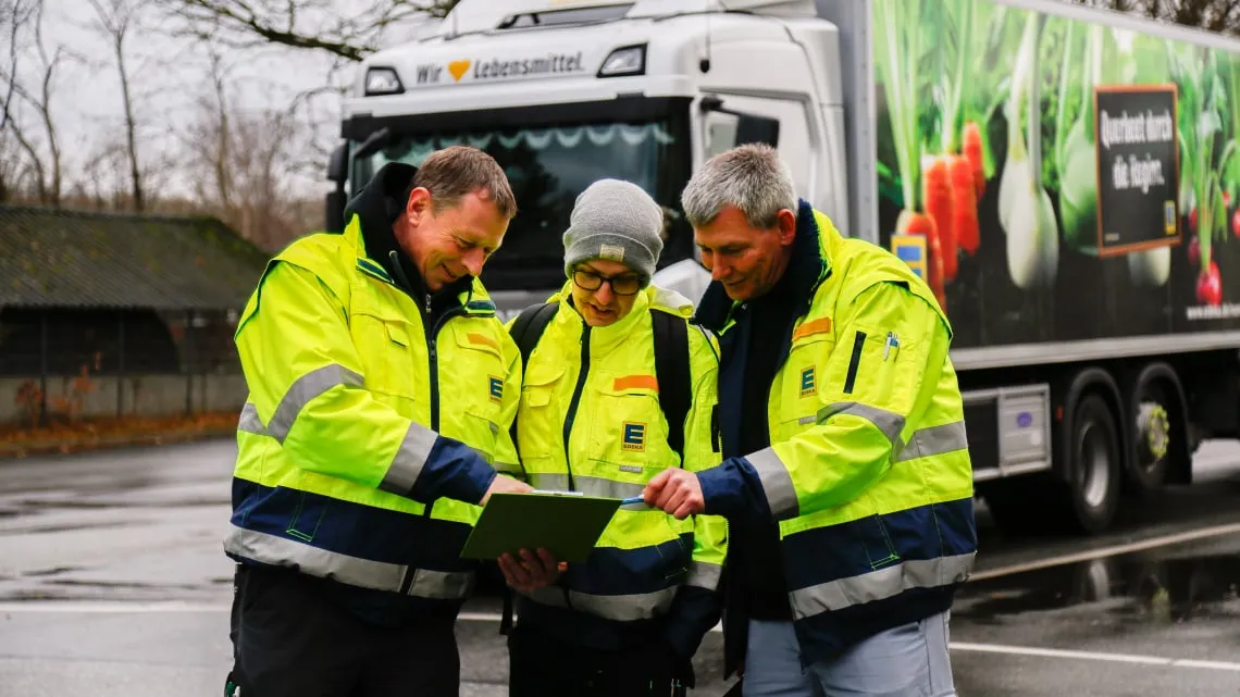 Drei EDEKA-Mitarbeitende in Warnjacken besprechen gemeinsam eine Liste vor einem LKW auf dem Firmengelände.