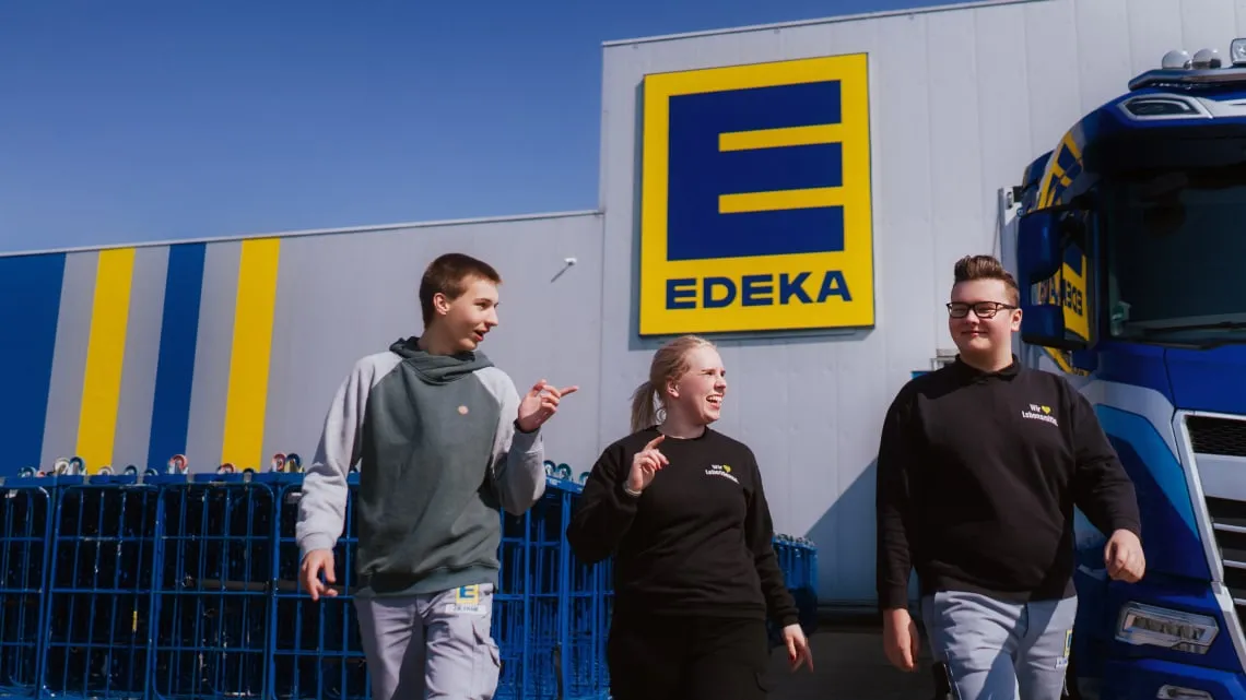 Drei junge Menschen lachen vor einem EDEKA-Markt und zeigen Dir einen freundlichen Einstieg ins Team.