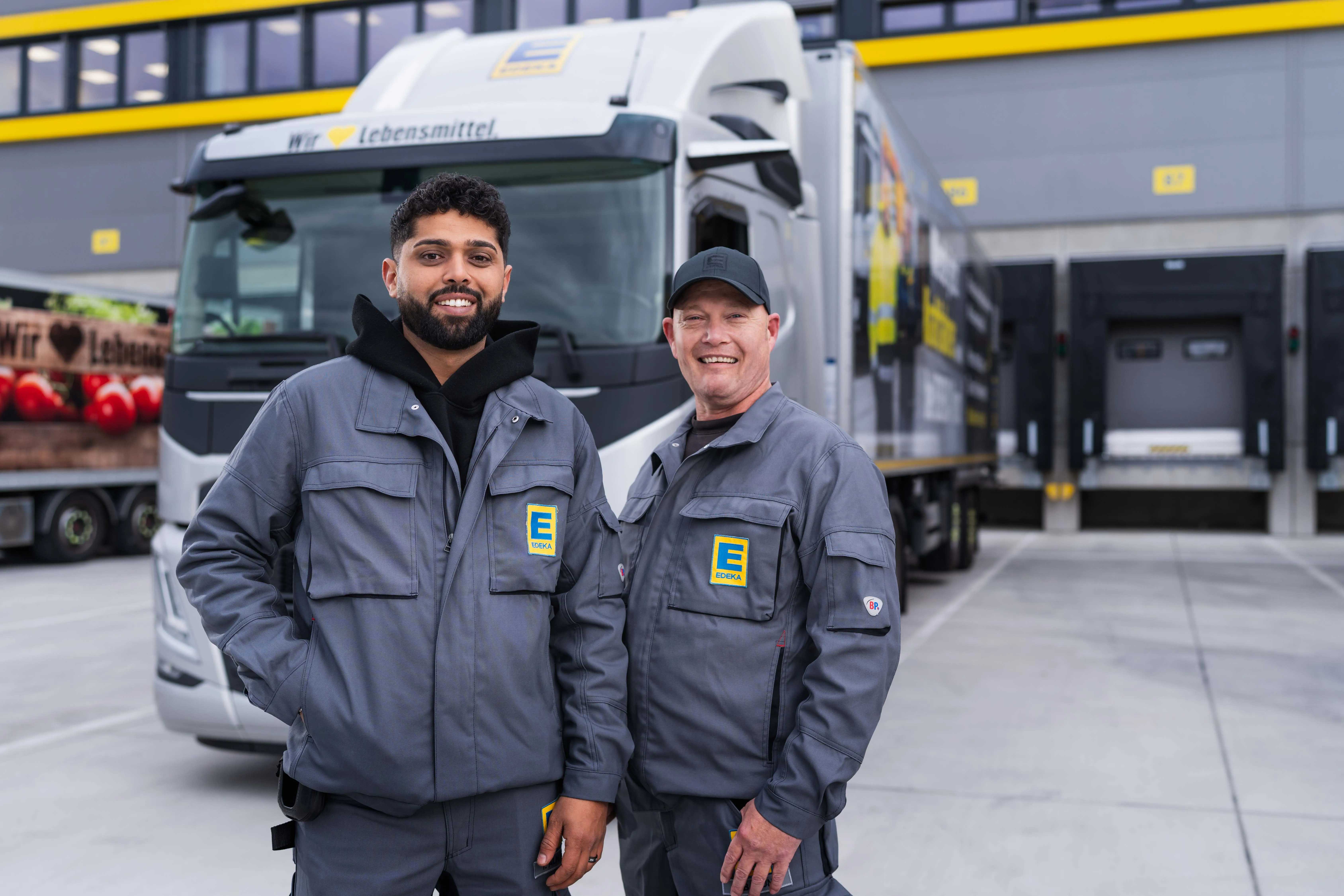 Zwei EDEKA-Mitarbeiter in Arbeitskleidung stehen lächelnd vor einem EDEKA-Lkw auf einem Firmengelände.
