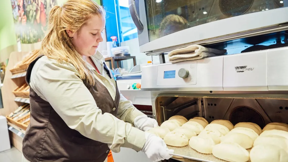 Du siehst eine Mitarbeiterin, die frische Brötchen in den Ofen eines EDEKA-Marktes schiebt.