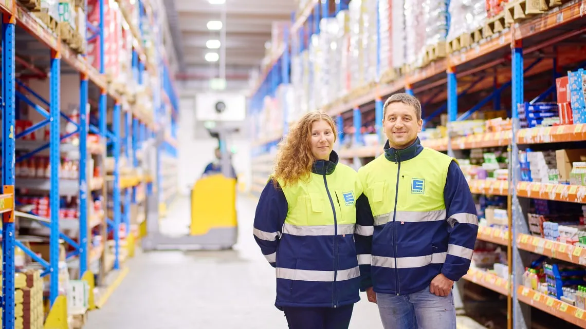 Zwei EDEKA-Mitarbeitende stehen lächelnd im Lager zwischen vollen Regalen und begrüßen Dich im Team.