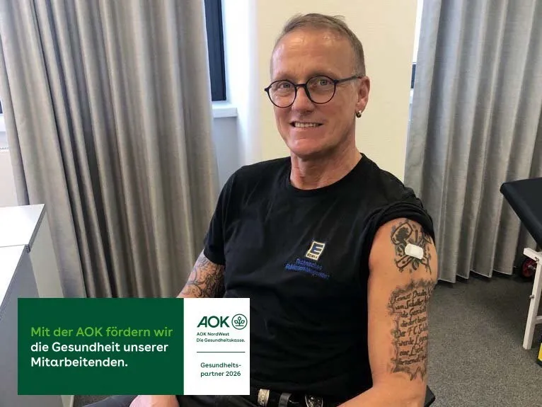 Ein lächelnder Mitarbeiter von EDEKA Nord zeigt stolz seine Tätowierungen. Ein AOK-Aufkleber betont die Gesundheitsförderung.