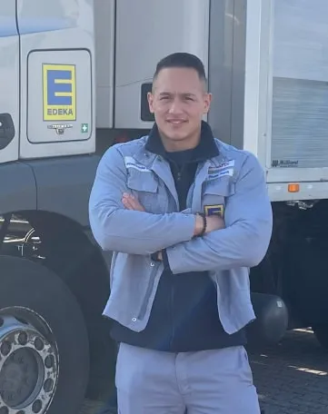 Ein junger EDEKA-Mitarbeiter steht mit verschränkten Armen vor einem EDEKA-Lkw und lächelt in die Kamera.
